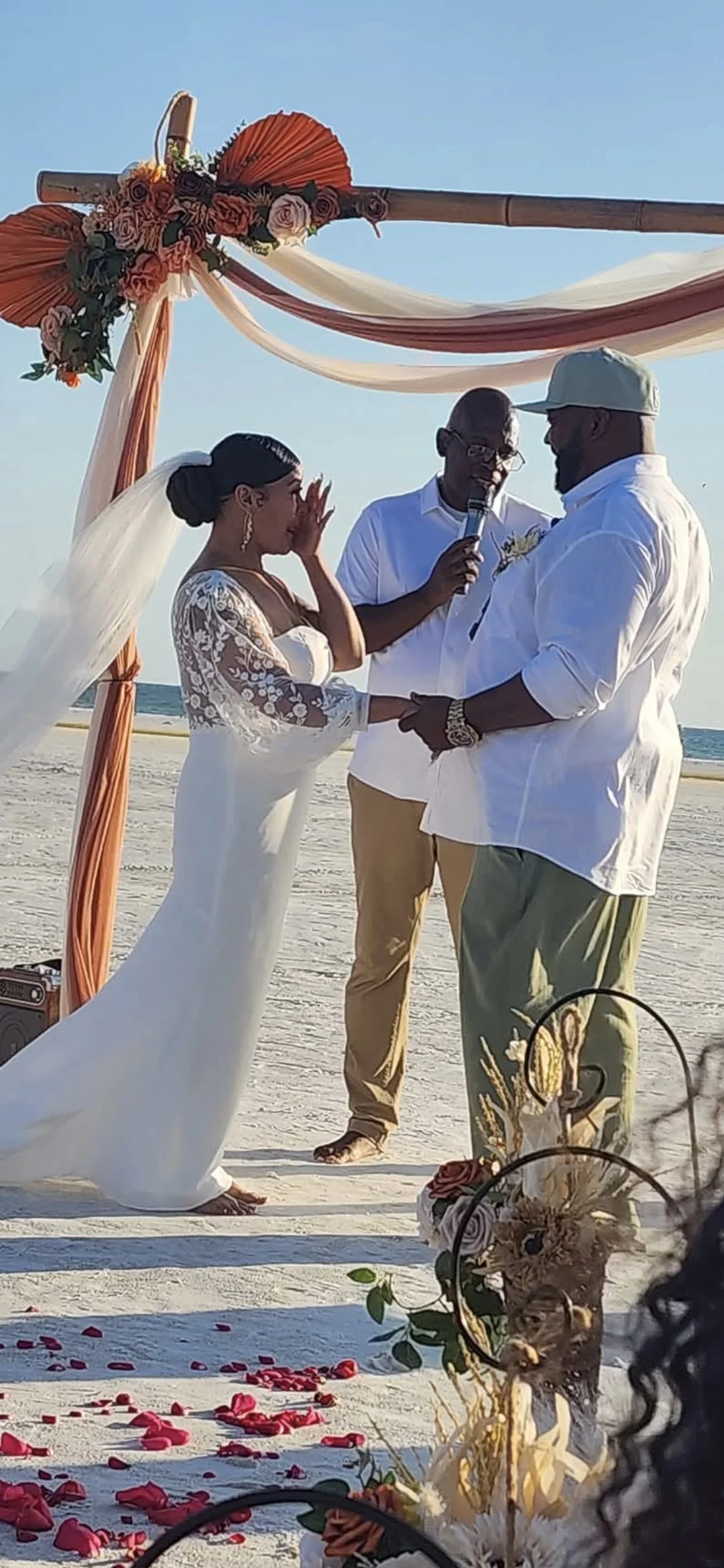 A beach wedding ceremony outdoors under a decorated arch with pink and white fabric and flowers. The bride and groom are holding hands, with the officiant reading from a microphone. Scattered rose petals are on the sand in the foreground.