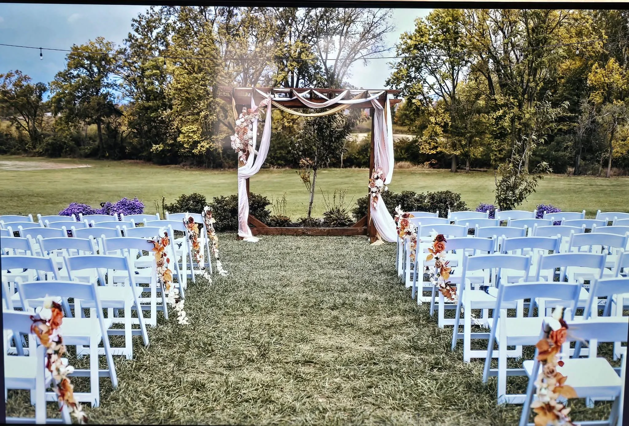 Outdoor wedding ceremony setup with white chairs decorated with flowers, an arch draped with white fabric, set on a grassy field with trees and string lights overhead.