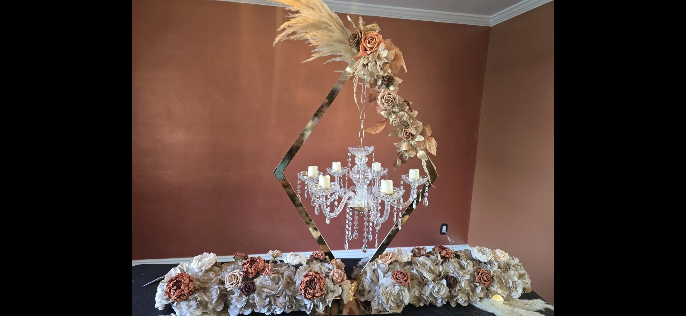 Decorative floral display featuring a crystal chandelier, arranged with pink, beige, and white roses, and pampas grass, set against a peach-colored wall.