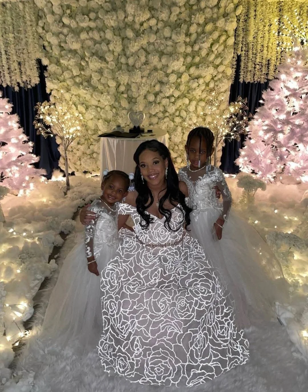 A woman and two young girls in white dresses posing together in a decorated indoor setting with white flowers, pink Christmas trees, and warm lighting.