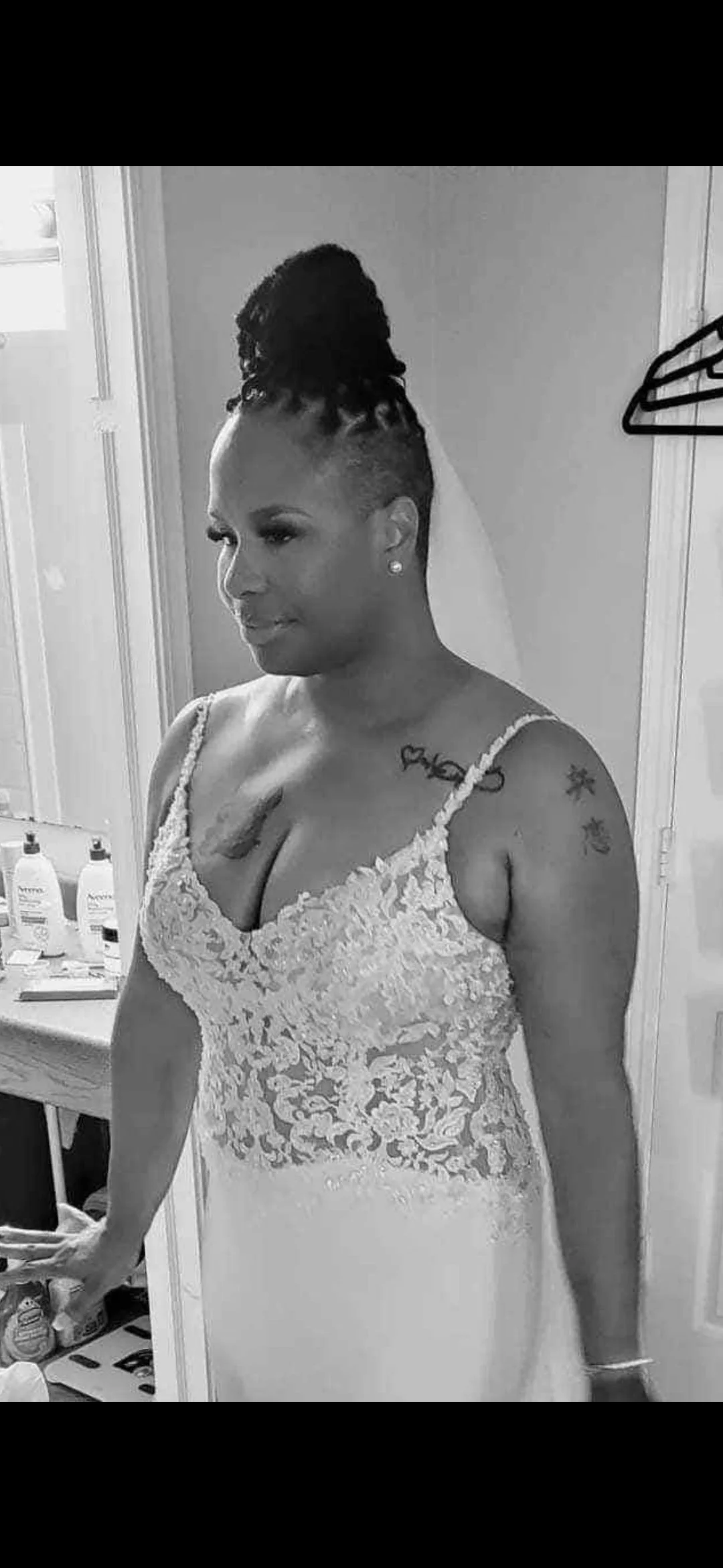 A woman in a wedding dress with lace details, standing in a room with various toiletries on a counter in the background.