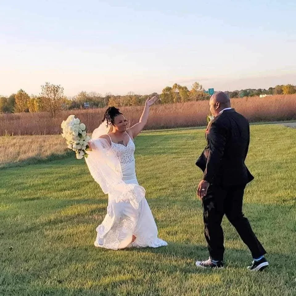 A bride in a wedding dress holding a bouquet is dancing on a grassy field with a man in a suit, under a clear sky.