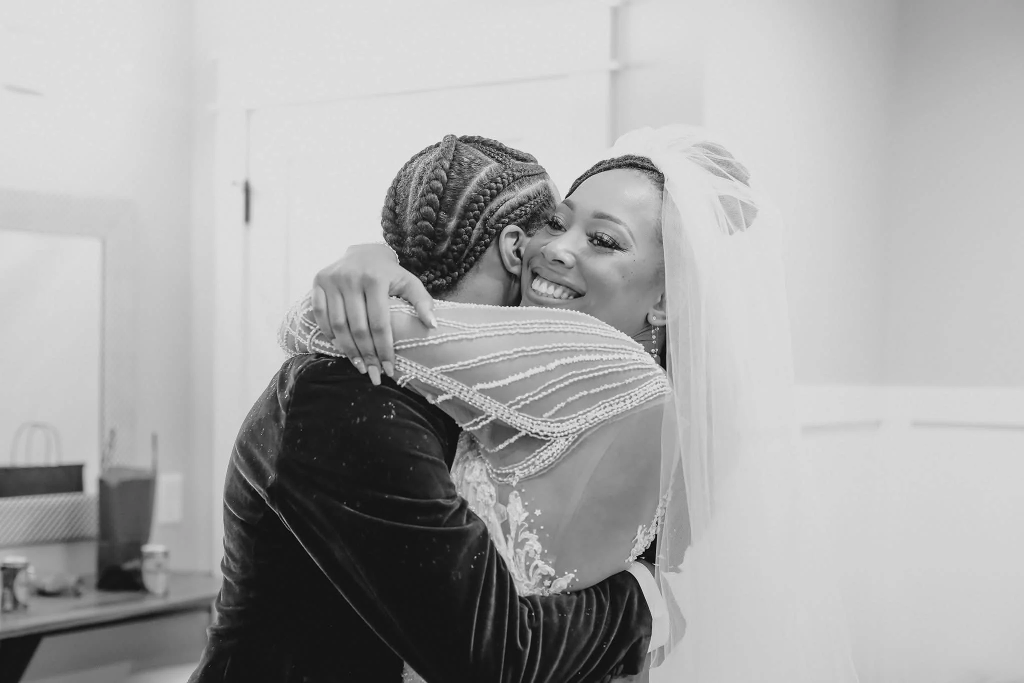 A woman in wedding attire and veil hugging a young girl with braided hair, smiling happily.