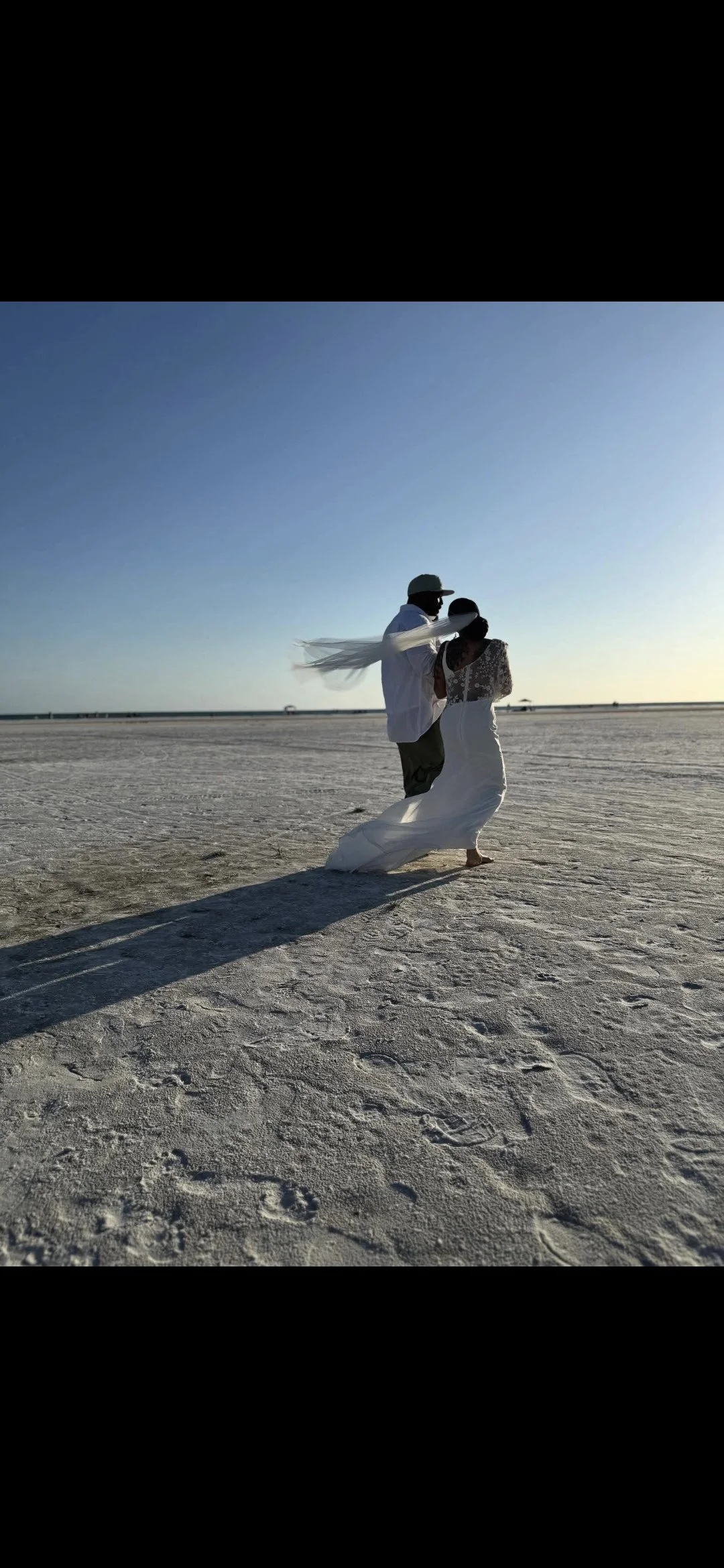 A couple dressed in wedding attire dancing on a sandy beach during sunset with long shadows cast on the sand.
