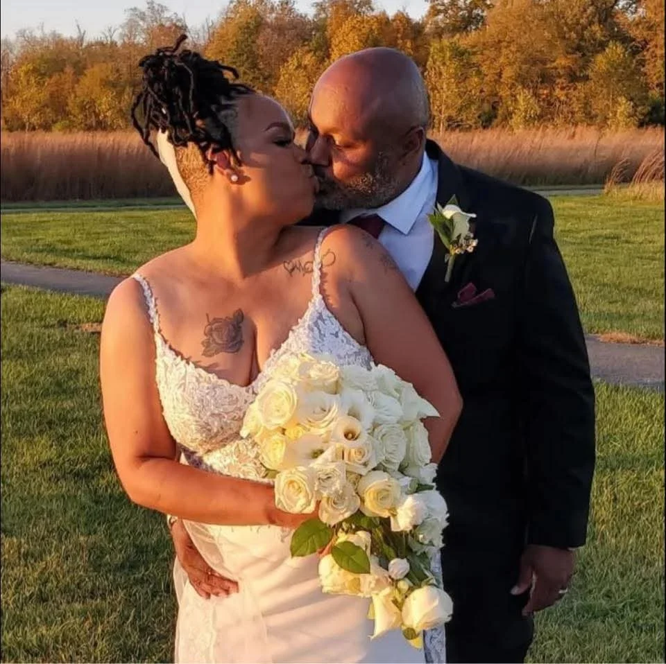 A bride and groom sharing a kiss outdoors during sunset, with the bride holding a large bouquet of white roses, wearing a lace wedding dress, and the groom in a black suit with a boutonniere.