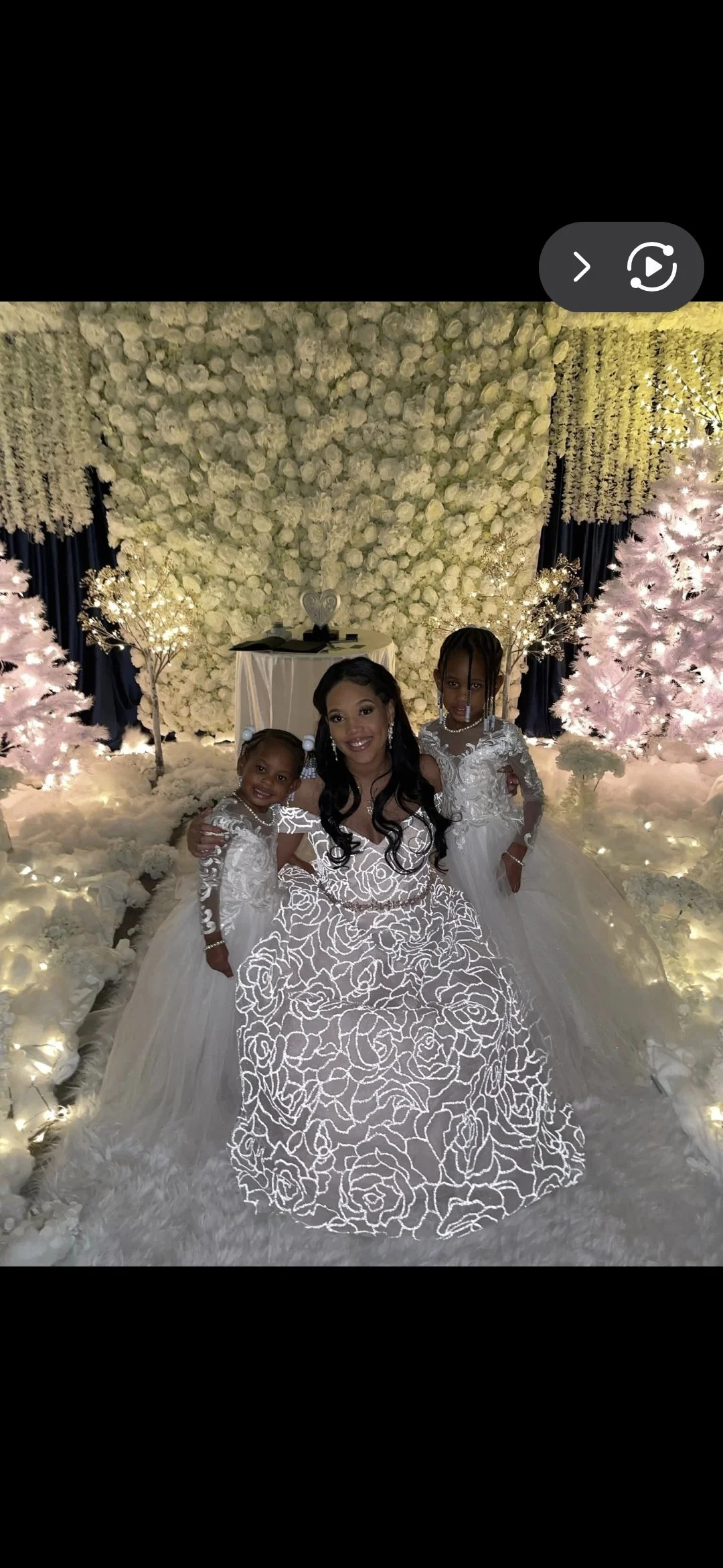 A woman in a white floral dress sitting on a white floral-covered bench, flanked by two young girls in white dresses, all smiling in a decorated indoor setting with white flowers, trees, and soft lighting.