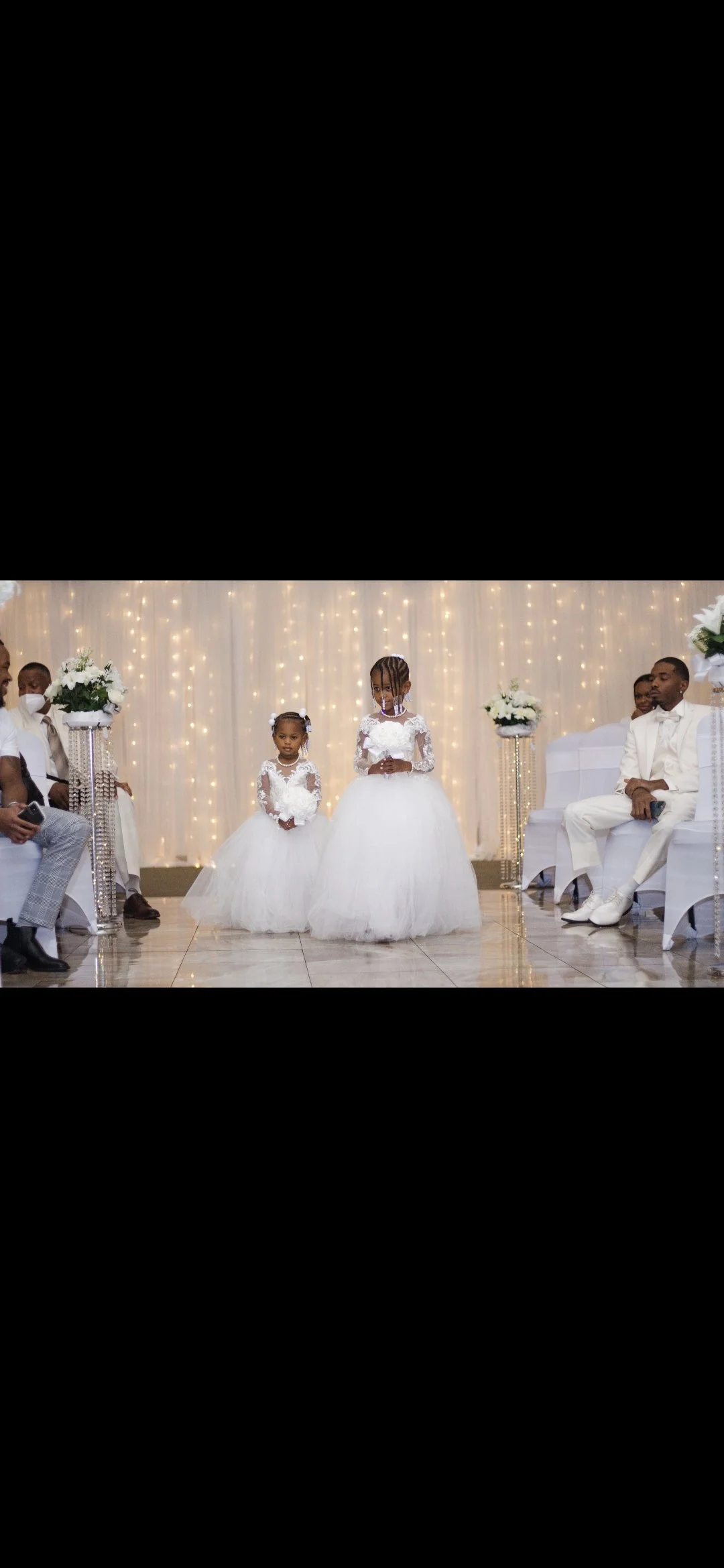 A wedding ceremony with two young girls dressed in white dresses standing in front of a decorated backdrop with string lights. Guests are seated on either side, some wearing masks.