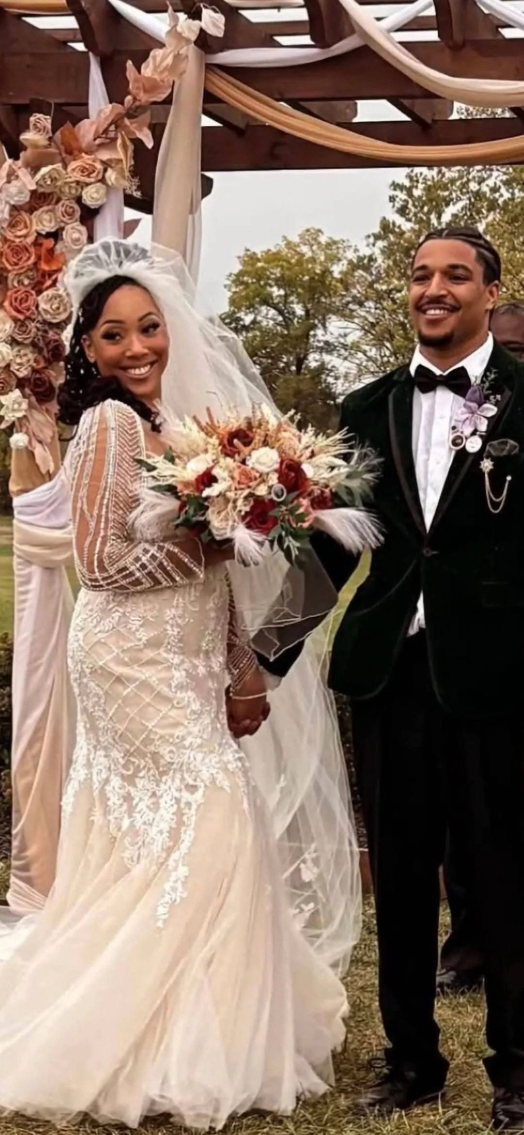 A bride and groom hold hands and smile at their wedding ceremony outdoors; the bride wears a lace wedding dress, and the groom wears a black tuxedo with a bow tie; the bride holds a bouquet of flowers.