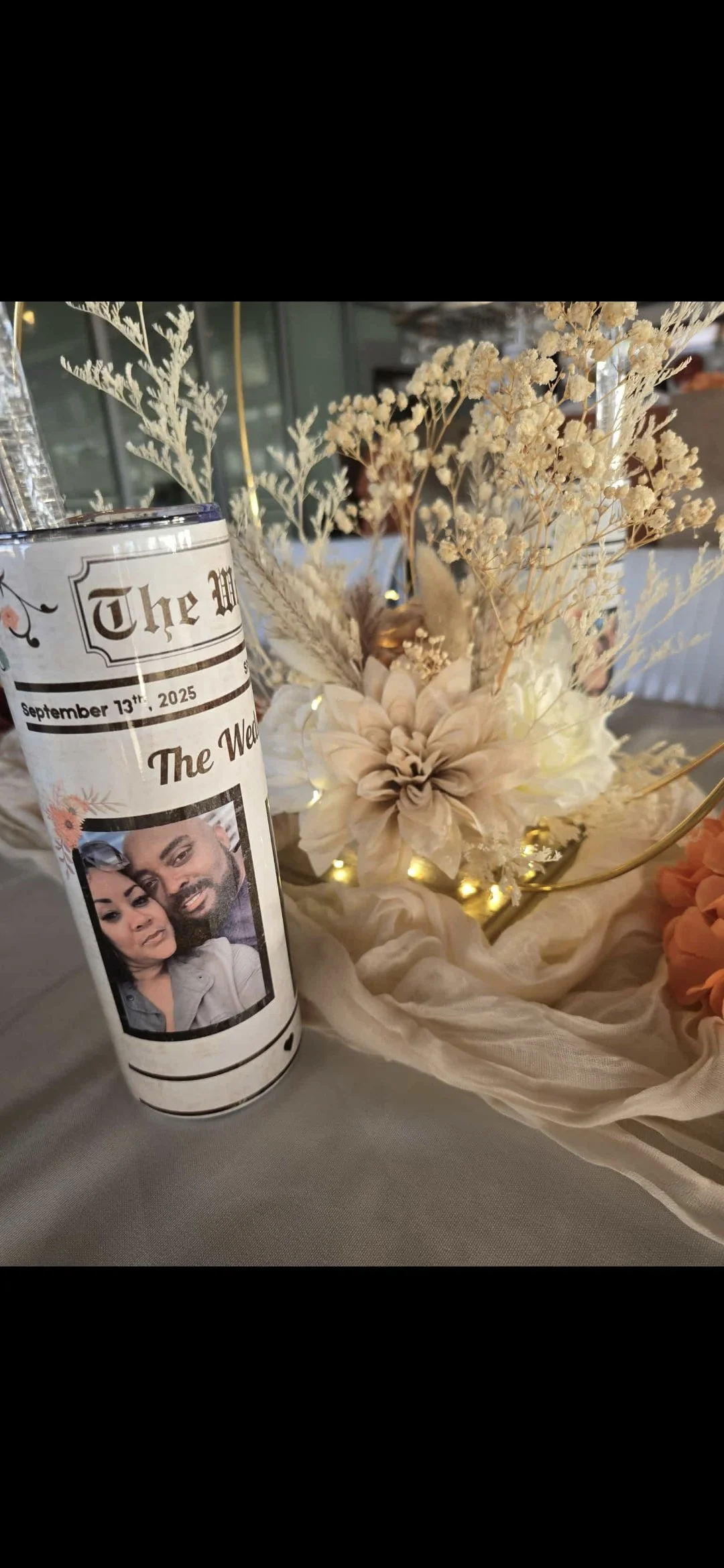 A floral centerpiece with dried white and beige flowers and greenery, illuminated by small string lights, beside a can decorated with a newsprint design and a photo of a couple, on a table at a wedding reception.