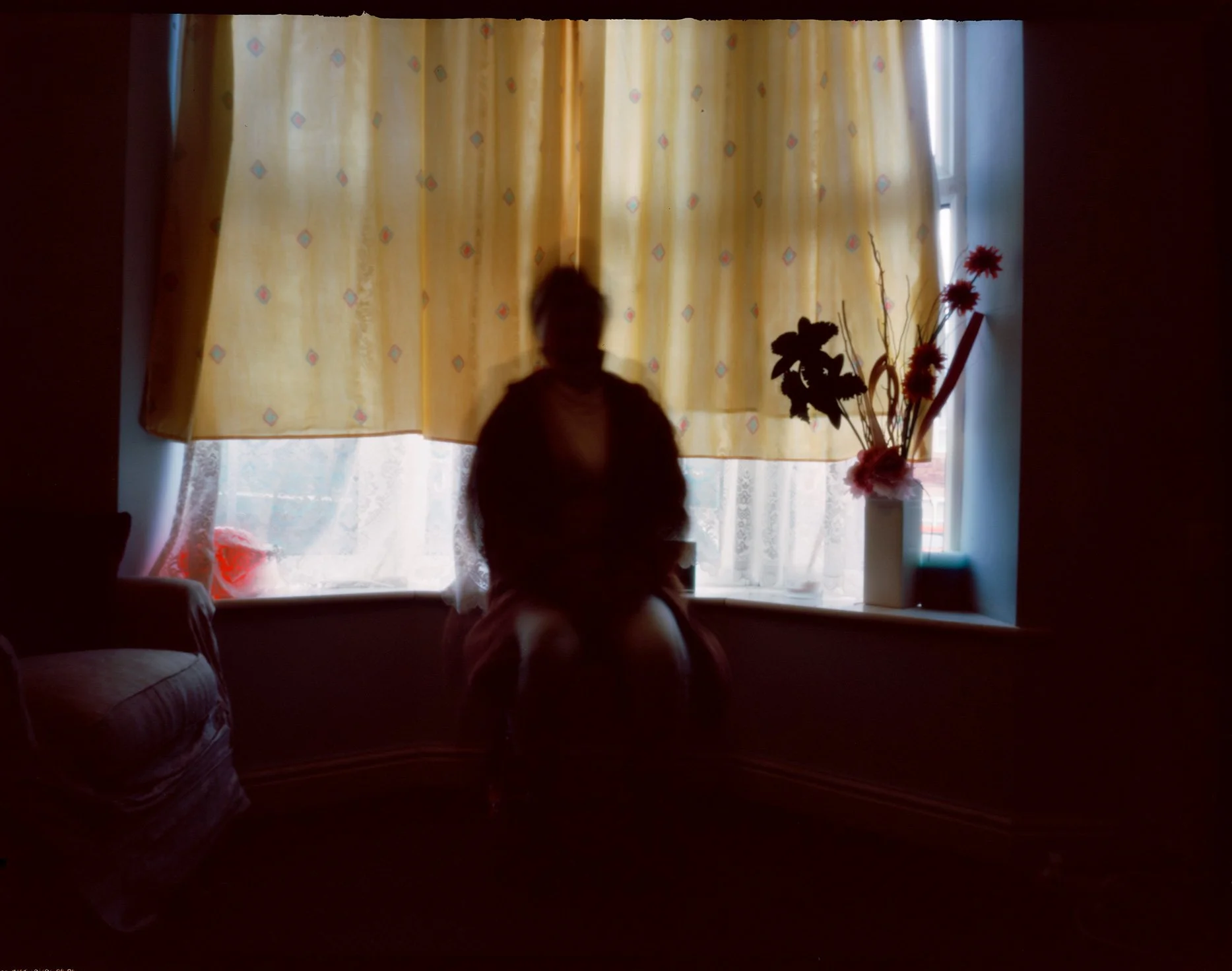 Silhouette of a person sitting by a window with yellow curtains, with a vase of flowers on the windowsill, backlit by daylight.