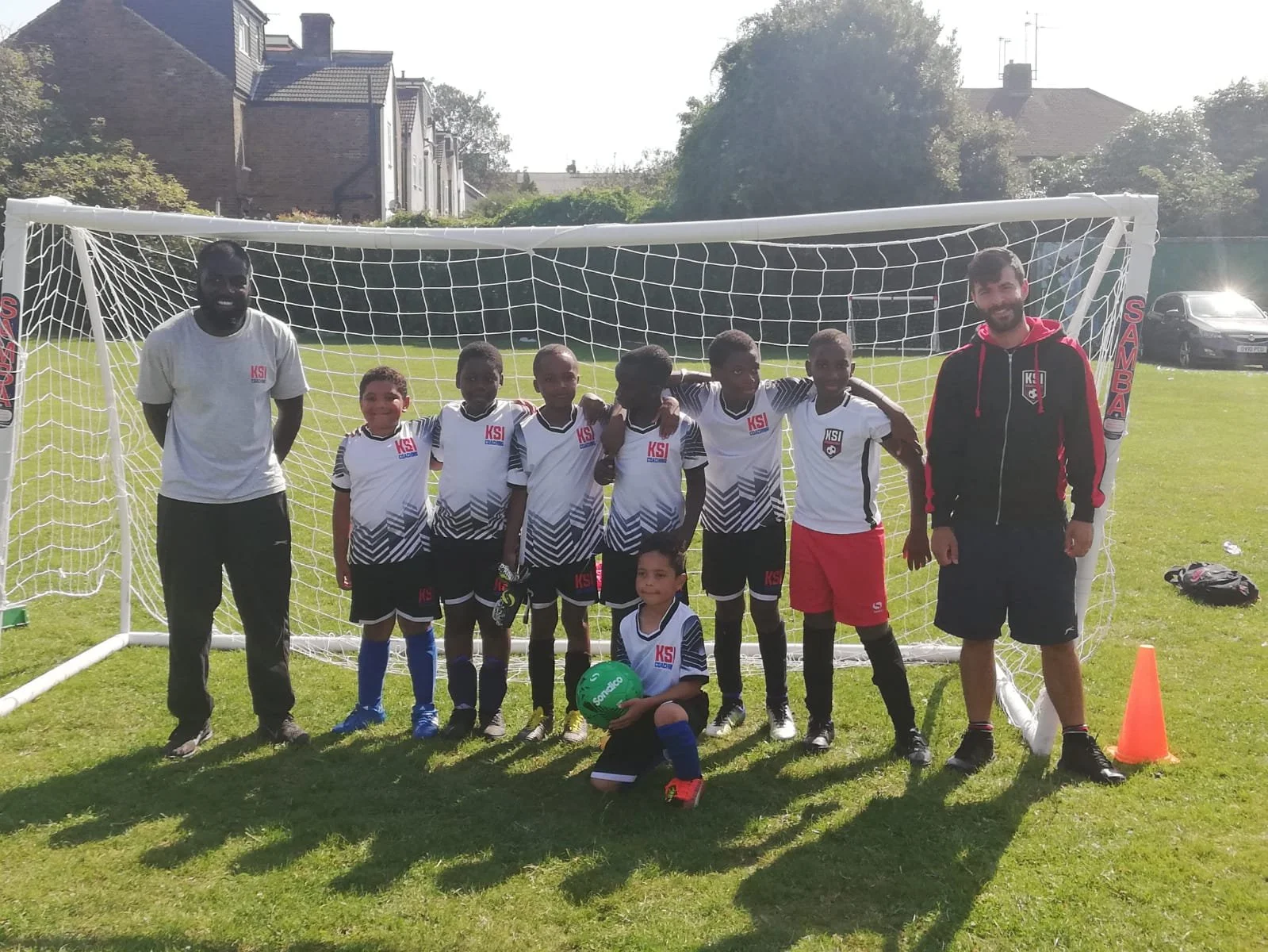 Group of young soccer players and two coaches lying and standing in front of a soccer goal on a grassy field on a sunny day.