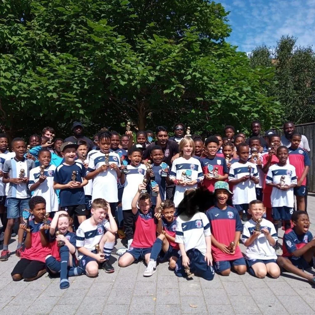 A group of children in sports jerseys, holding trophies, smiling and posing for a photo outdoors on a sunny day under a large leafy tree.