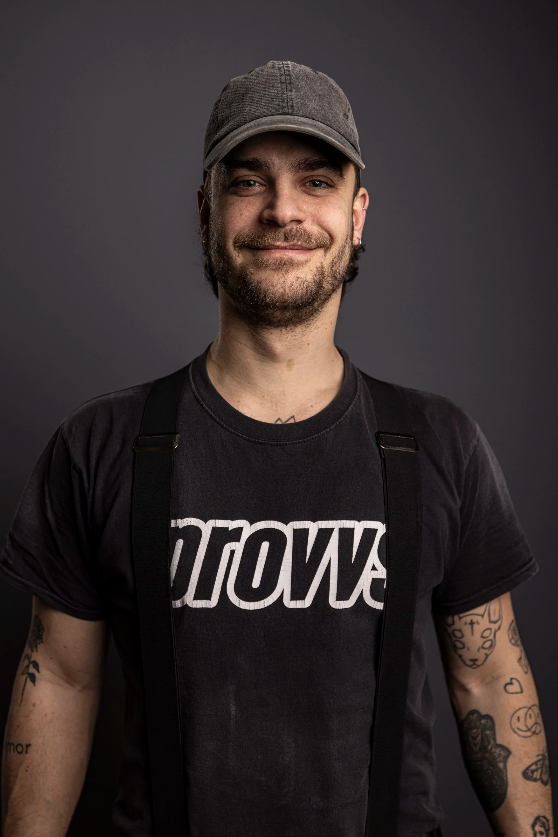 En ung man med mörkt hår, skägg och tatueringar på armarna, bär en svart t-shirt med texten 'SNOW' och en grå keps, poserar mot en mörk bakgrund.