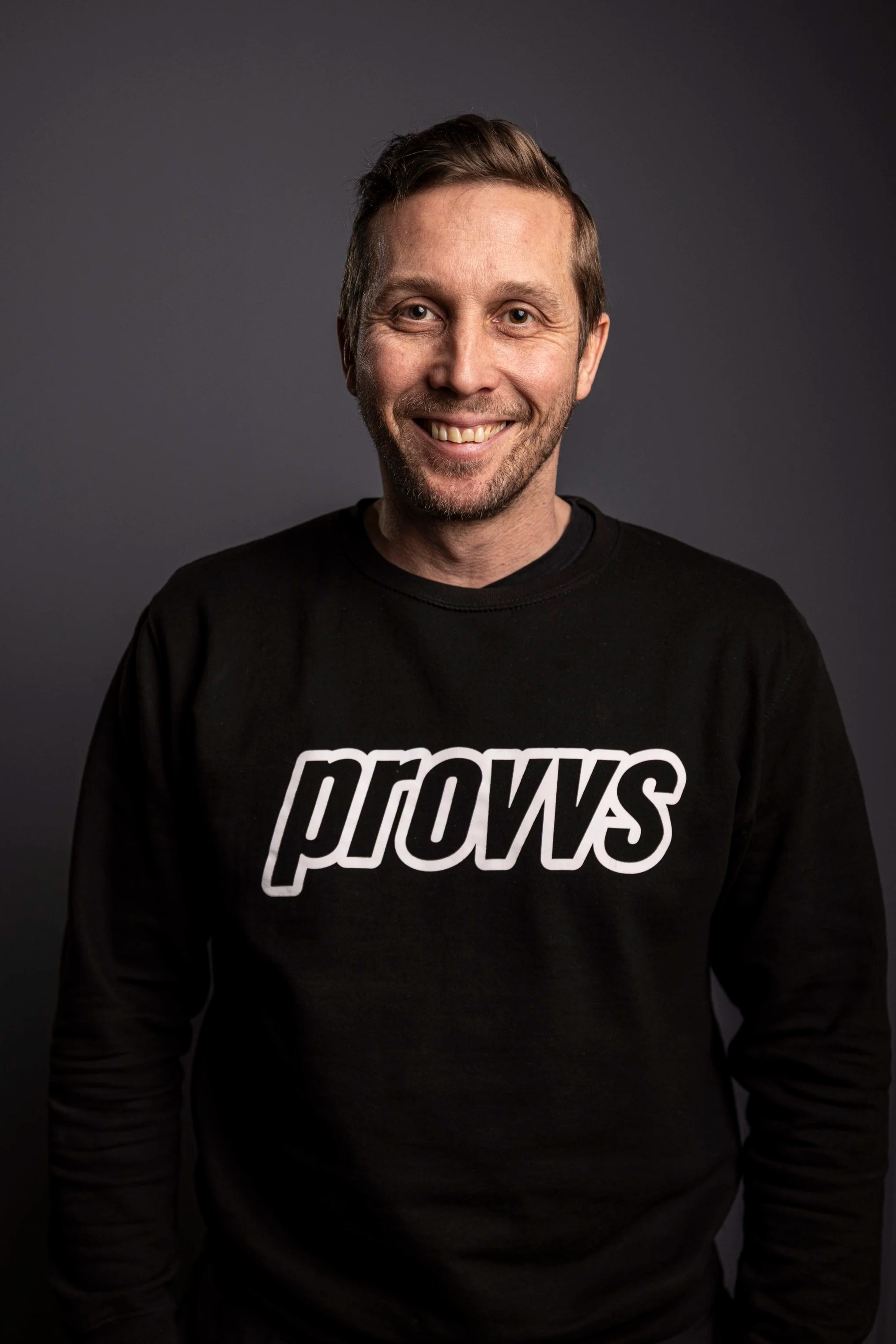 En man med kort hår, skägg och ett leende, bär en svart tröja med texten 'prows' på.
