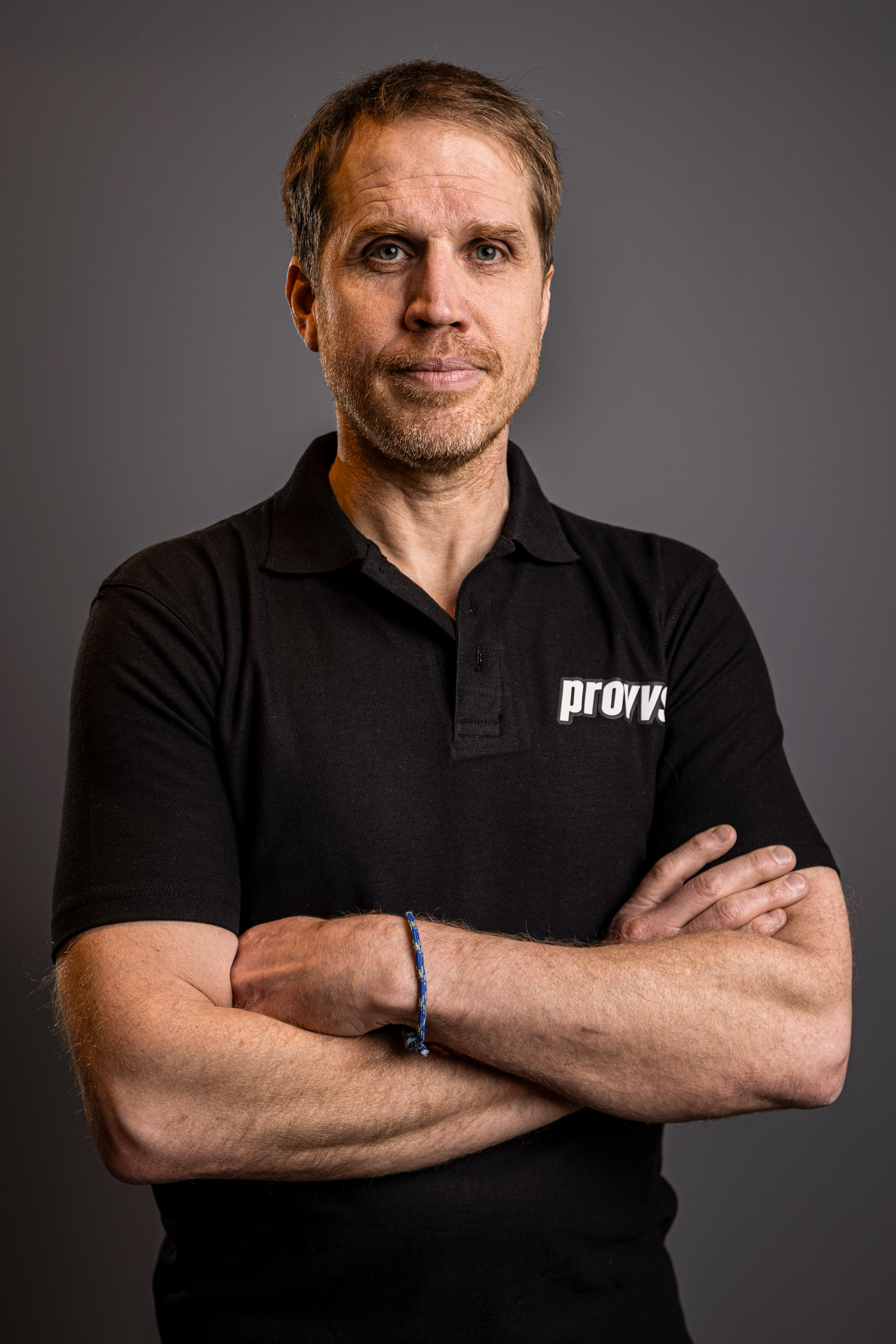 En man med blont hår och skägg, korsade armar, bär en svart polo med 'prove' på bröstet, står mot en grå bakgrund.