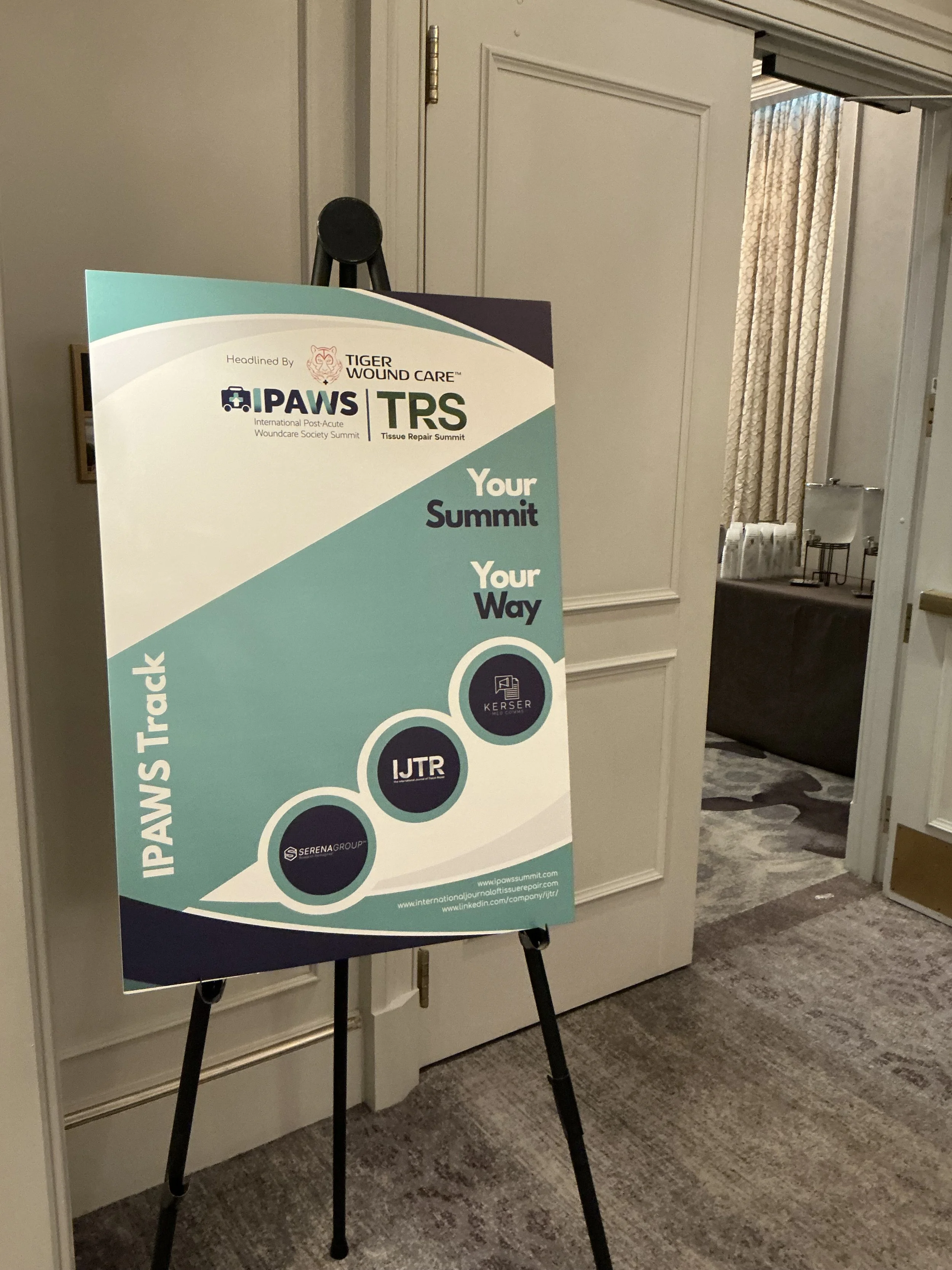 Event poster for a medical summit on wound care, tissue repair, and post-acute wound management, featuring logos of organizations like IPWS, TIGER WOUND CARE, and TRS, displayed on an easel in a conference room.