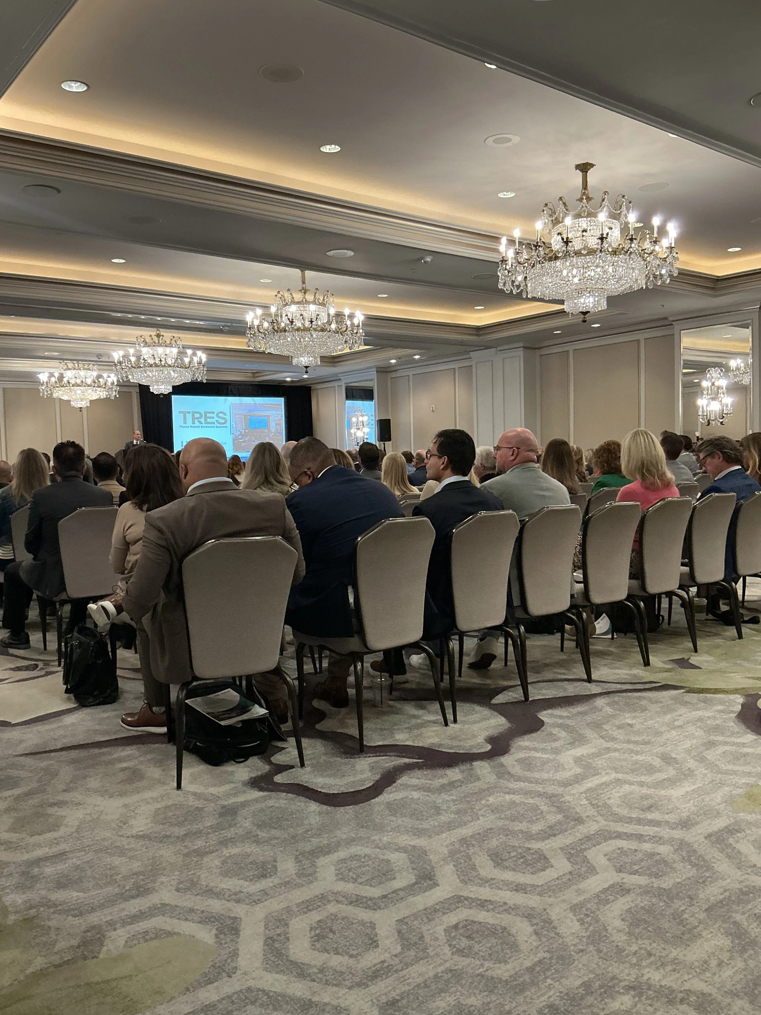 A large conference room filled with people seated in rows, watching a presentation on a screen at the front. The room has ornate chandeliers and elegant ceiling lighting.