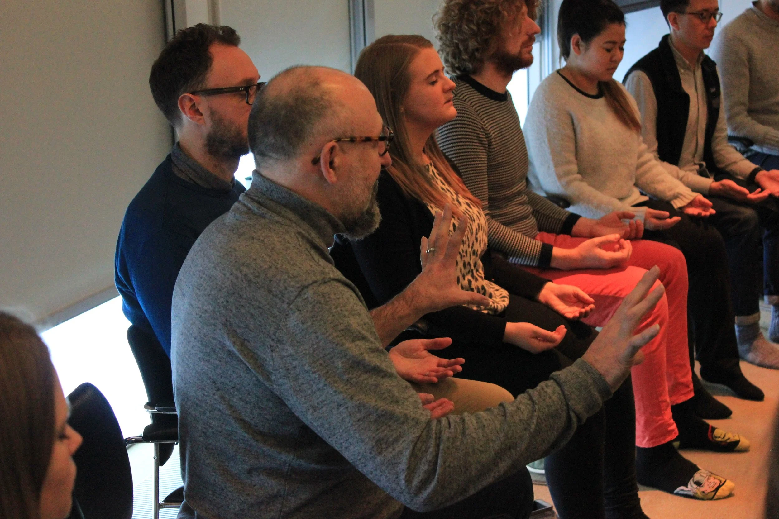 Group of people seated in a row during a meditation or mindfulness session, some with eyes closed and hands in a meditative pose, in a well-lit room.