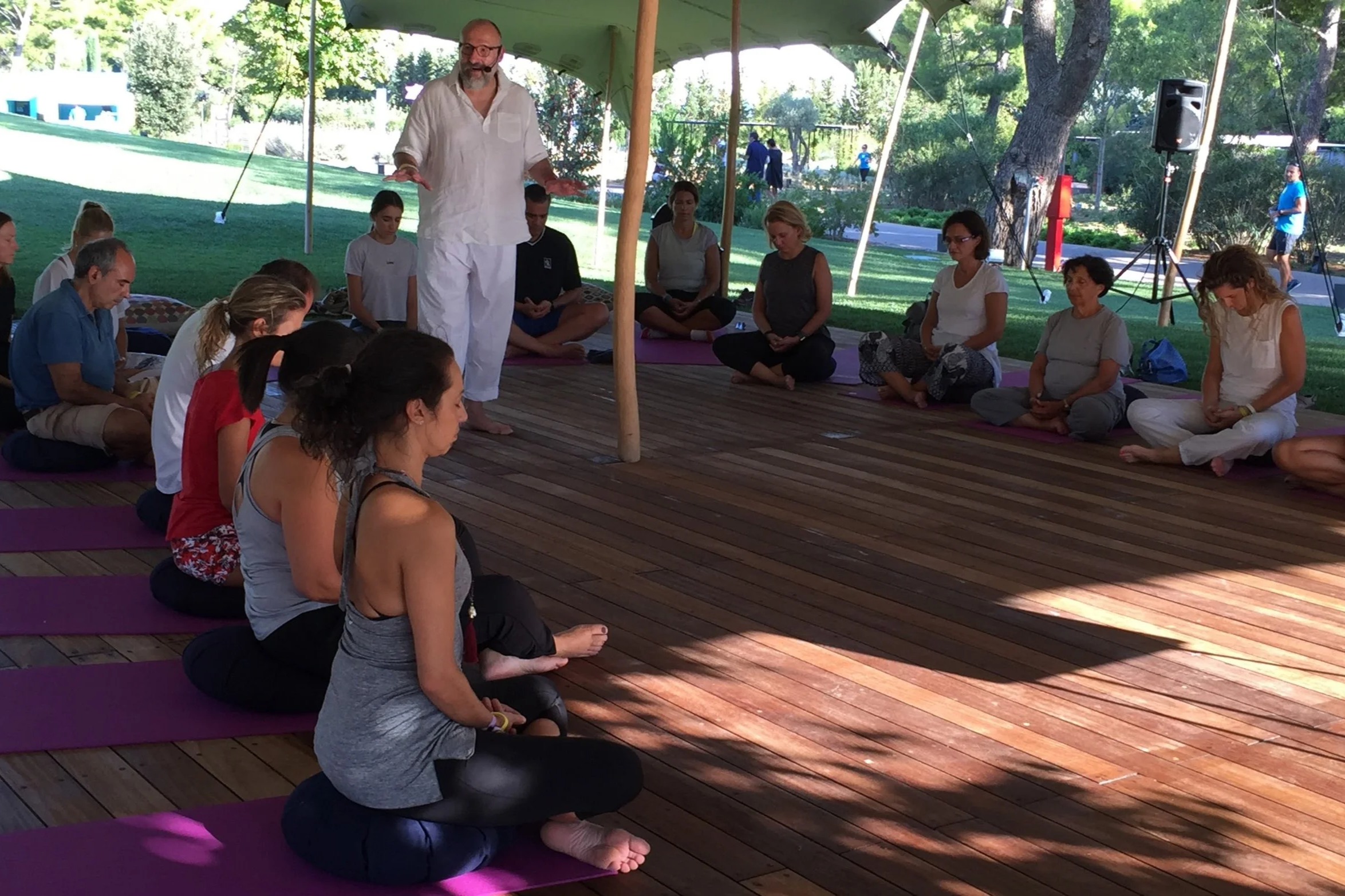 A group of people participating in a meditation or yoga session under a large tent outdoors, sitting cross-legged on purple mats, with a man in white leading the session.