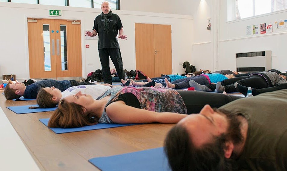 A group of people lying on exercise mats in a yoga or meditation class, with an instructor standing and gesturing in front of them, in a bright room with natural light.