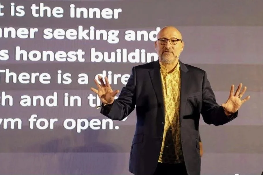 A man with glasses and a beard, wearing a dark blazer and a patterned yellow shirt, standing in front of a projection screen with white text. He is gesturing with both hands, palms facing outward.