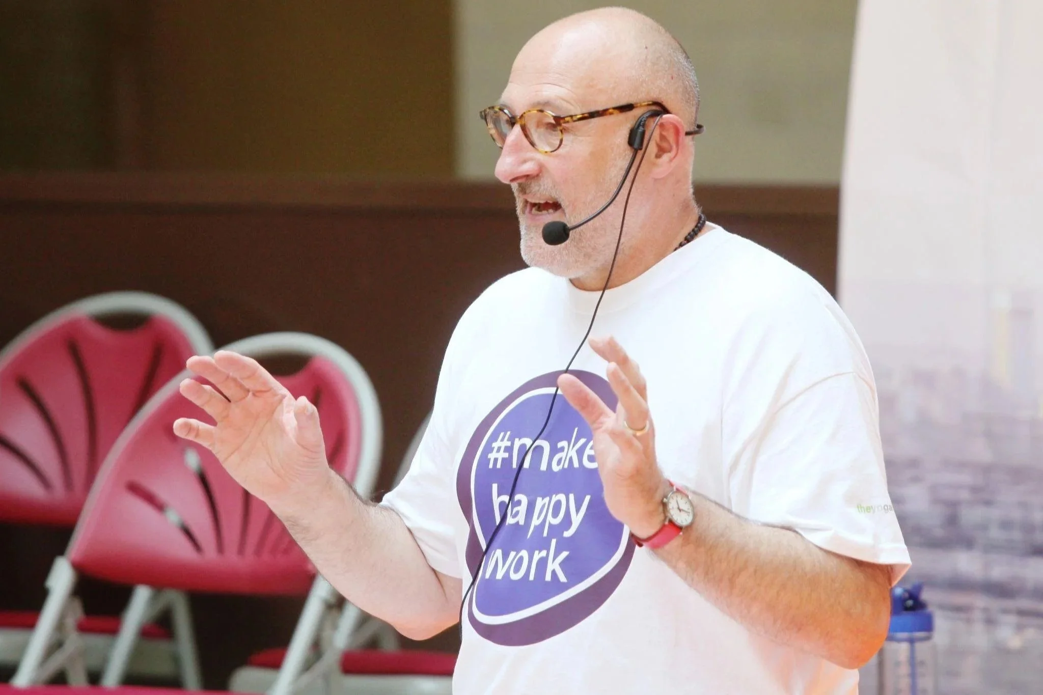 A man speaking at an event, wearing glasses, a headset microphone, and a white t-shirt with the hashtag '#makinghappywork'.