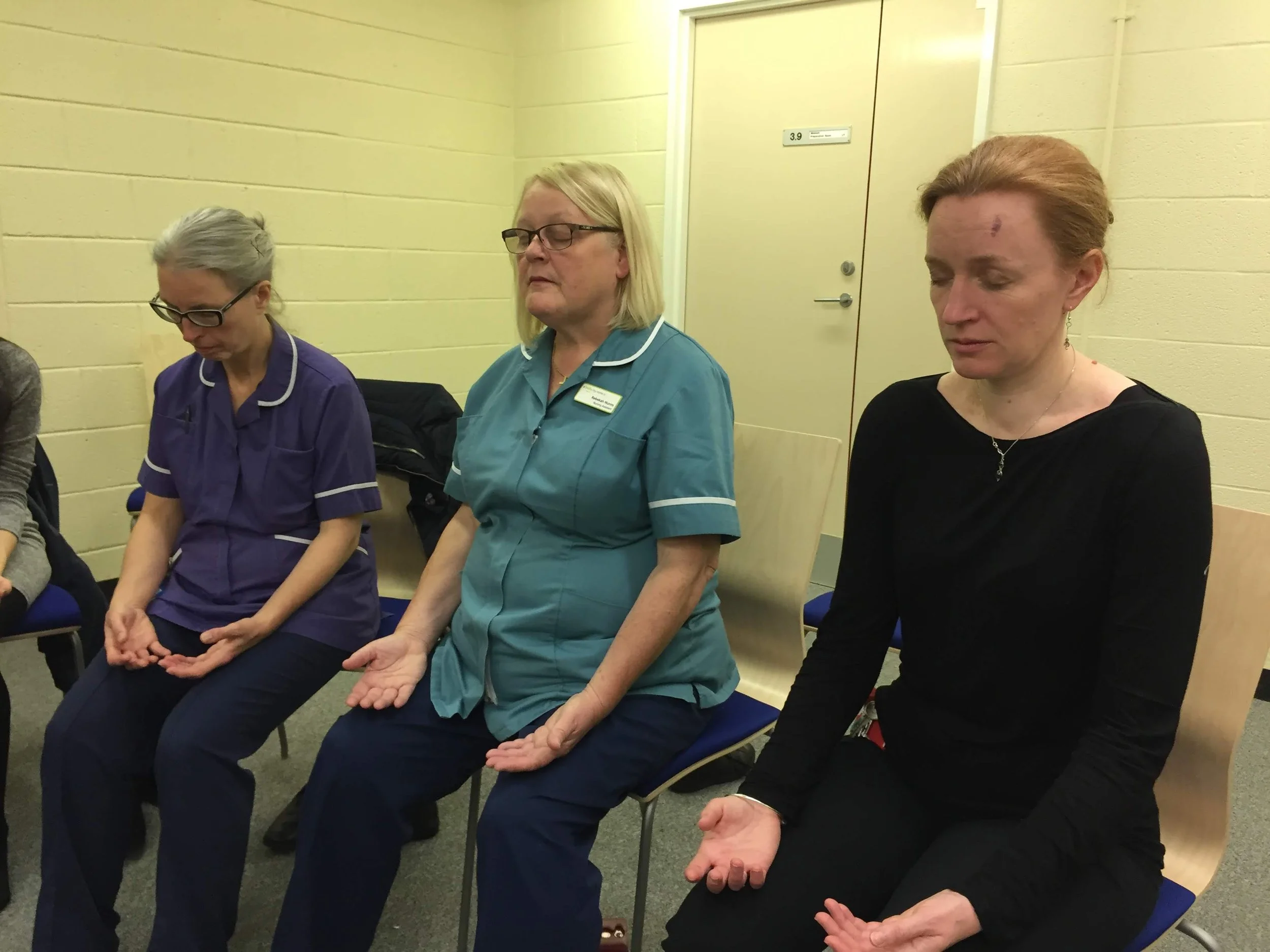 Three women sitting on chairs in a room, appears to be a prayer or meditation session, eyes closed, hands resting on laps or knees, with a yellow wall and a door in the background.