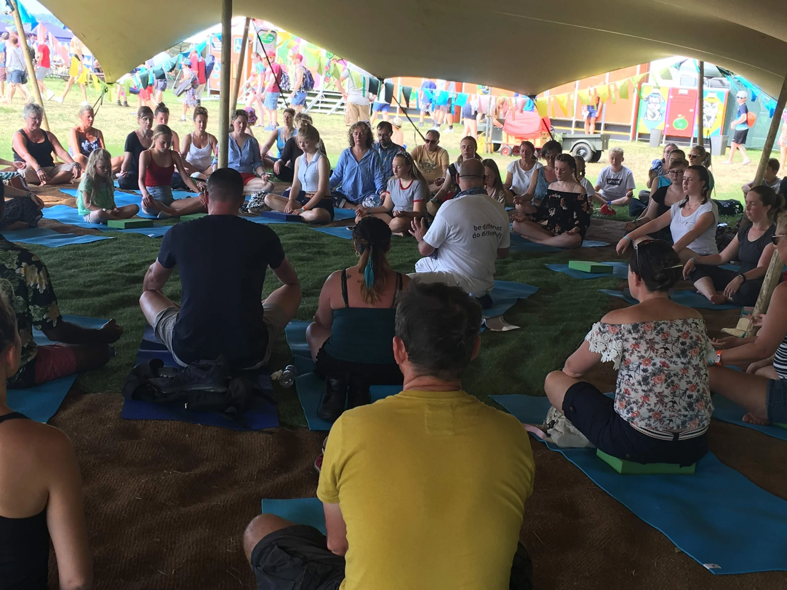 People participating in a yoga or meditation class under a large tent at an outdoor festival, sitting cross-legged on mats, with colorful flags and stalls in the background.
