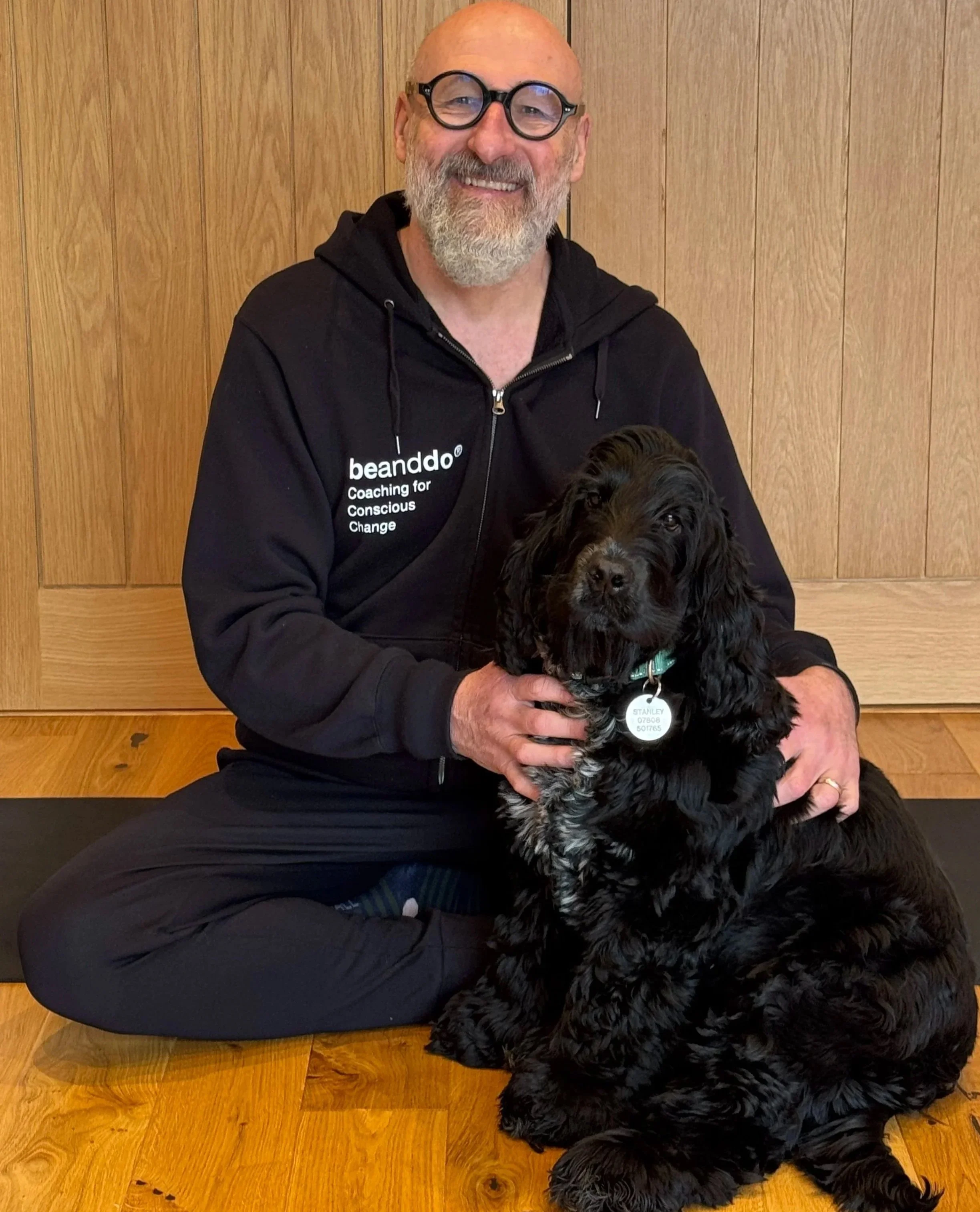 A man with glasses and a beard, wearing a black hoodie that says 'beanddo Coaching for Conscious Change,' is sitting cross-legged on a wooden floor, smiling at the camera, with a black and brown spaniel sitting in front of him. The man is holding the dog gently.