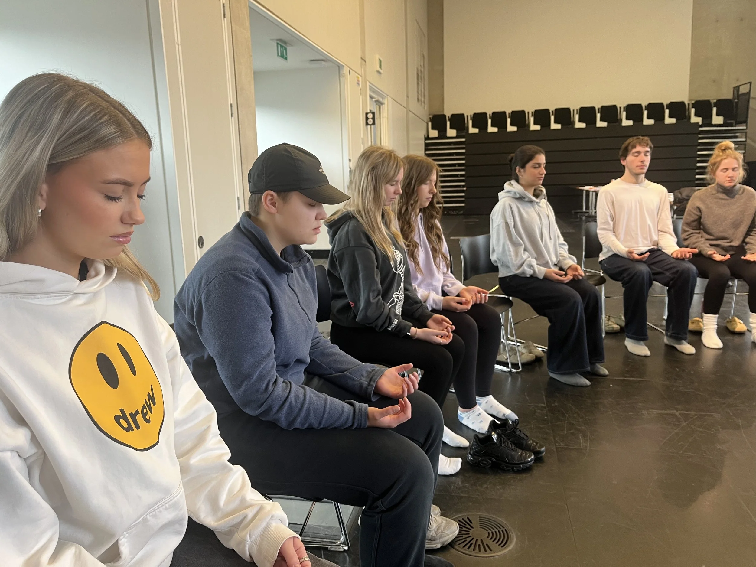 A group of young people seated in a circle with eyes closed, appearing to meditate or practice mindfulness in a studio or classroom.