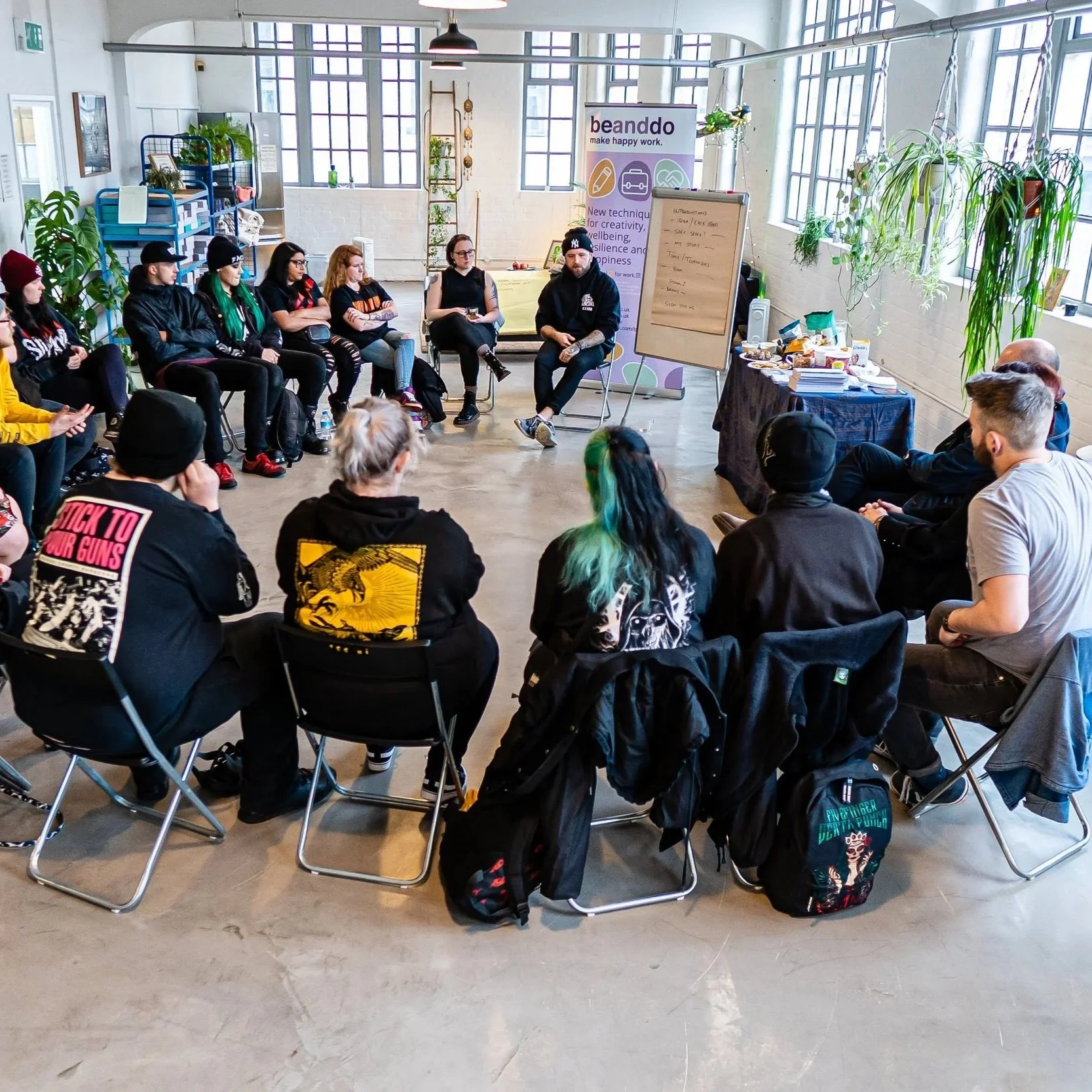 A group of young people participating in a workshop or discussion in a bright, industrial-style room with large windows and potted plants. They are sitting in a circular arrangement, with a facilitator at the front near a whiteboard and a banner that says 'beanddo.'