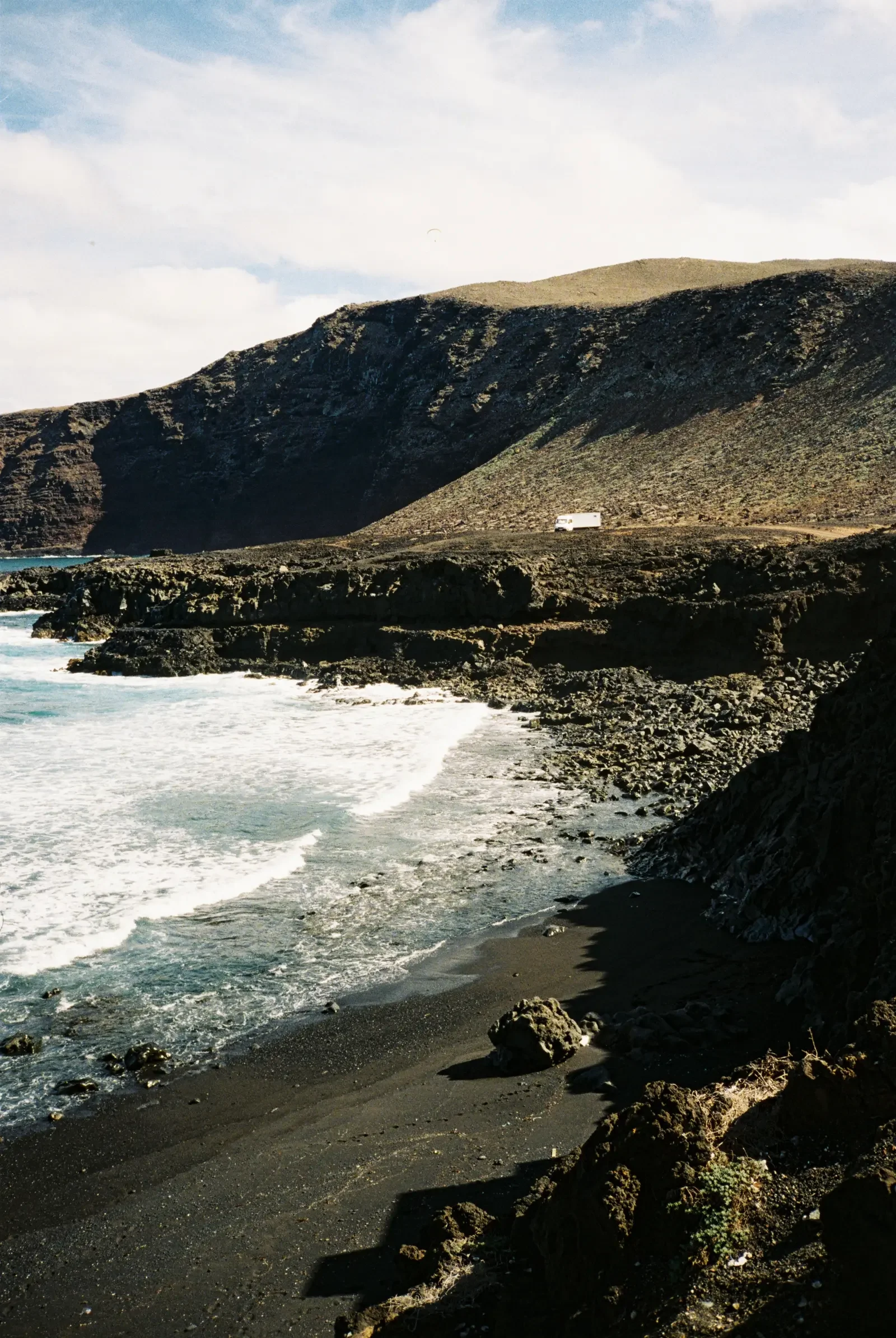 A rugged coastline with black sand beach, rocky cliffs, and waves crashing onto the shore. A mountain range is visible in the background, and a small white truck is parked near the base of the mountains.