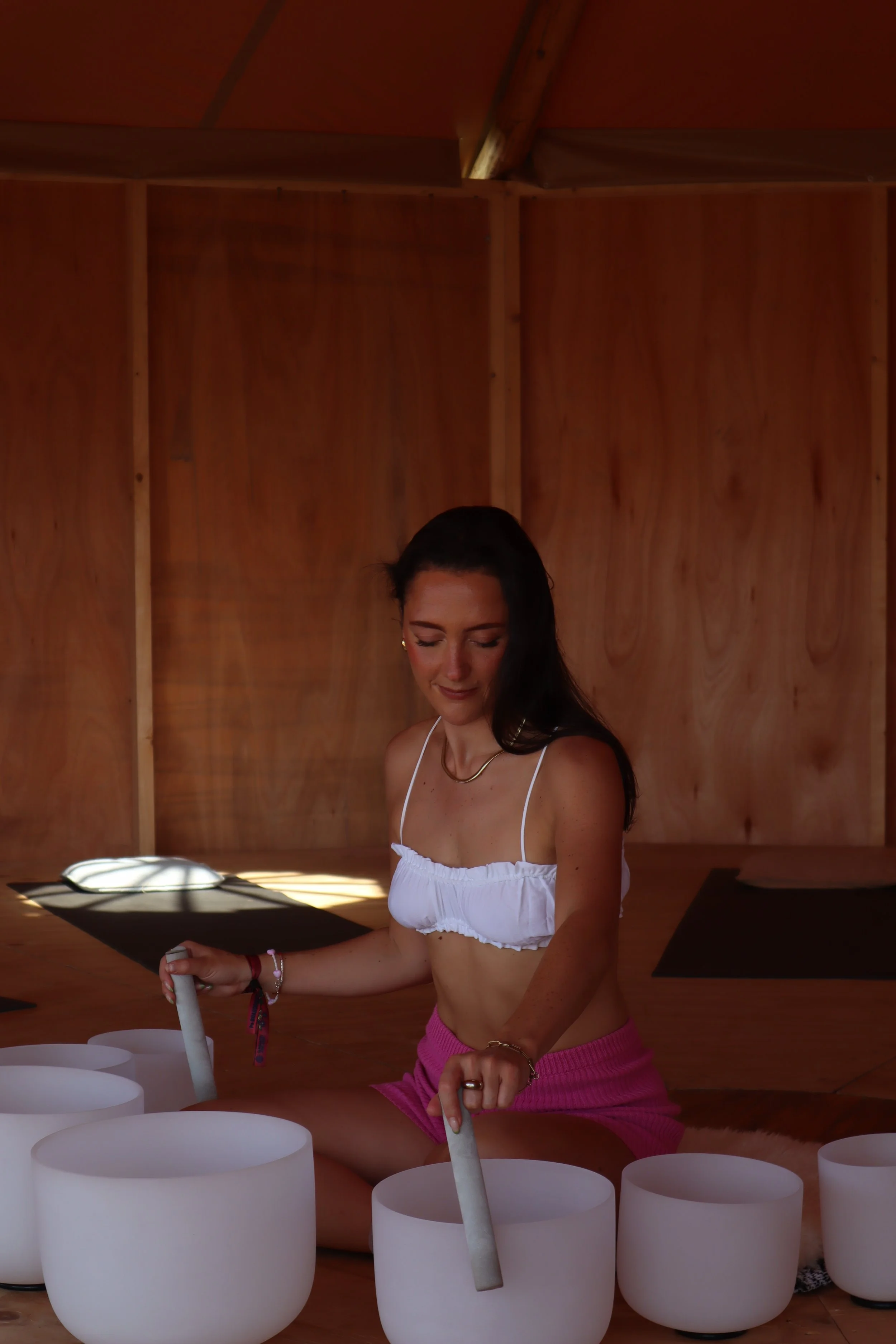 A woman sitting on the floor inside a wooden room, playing crystal singing bowls with mallets.