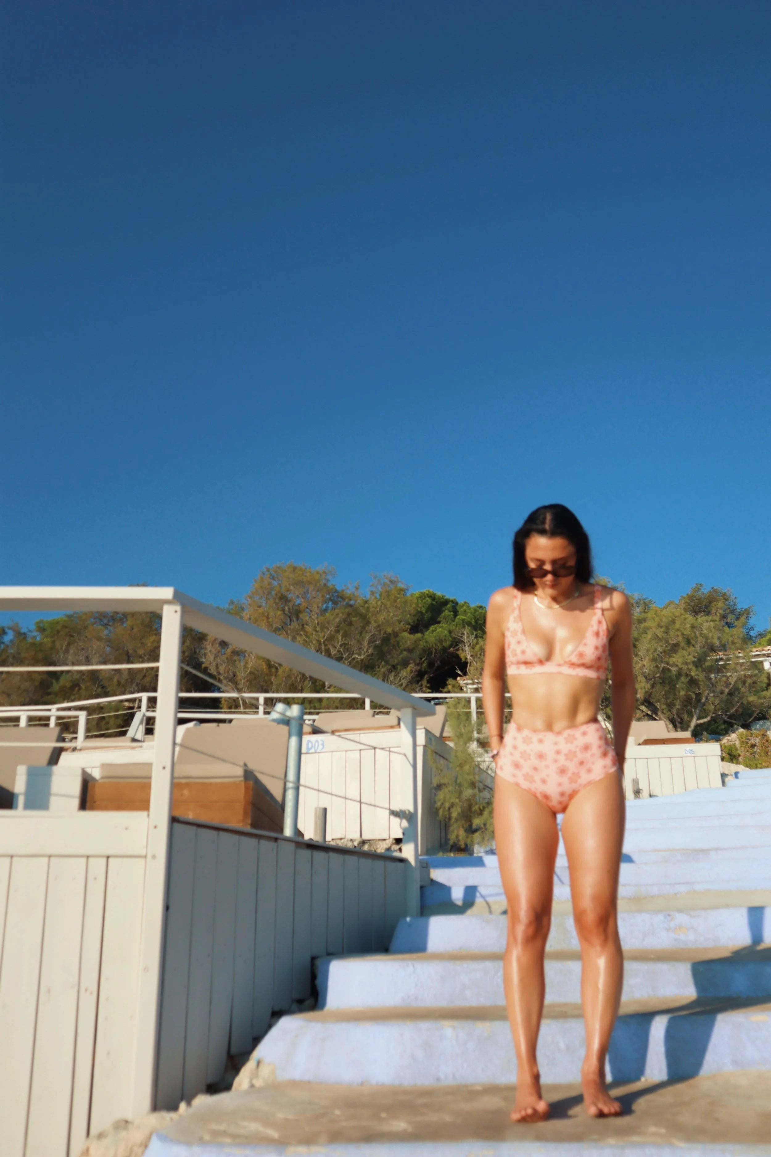 Woman in summer swimsuit walking down outdoor stairs on sunny day with blue sky and trees in background.