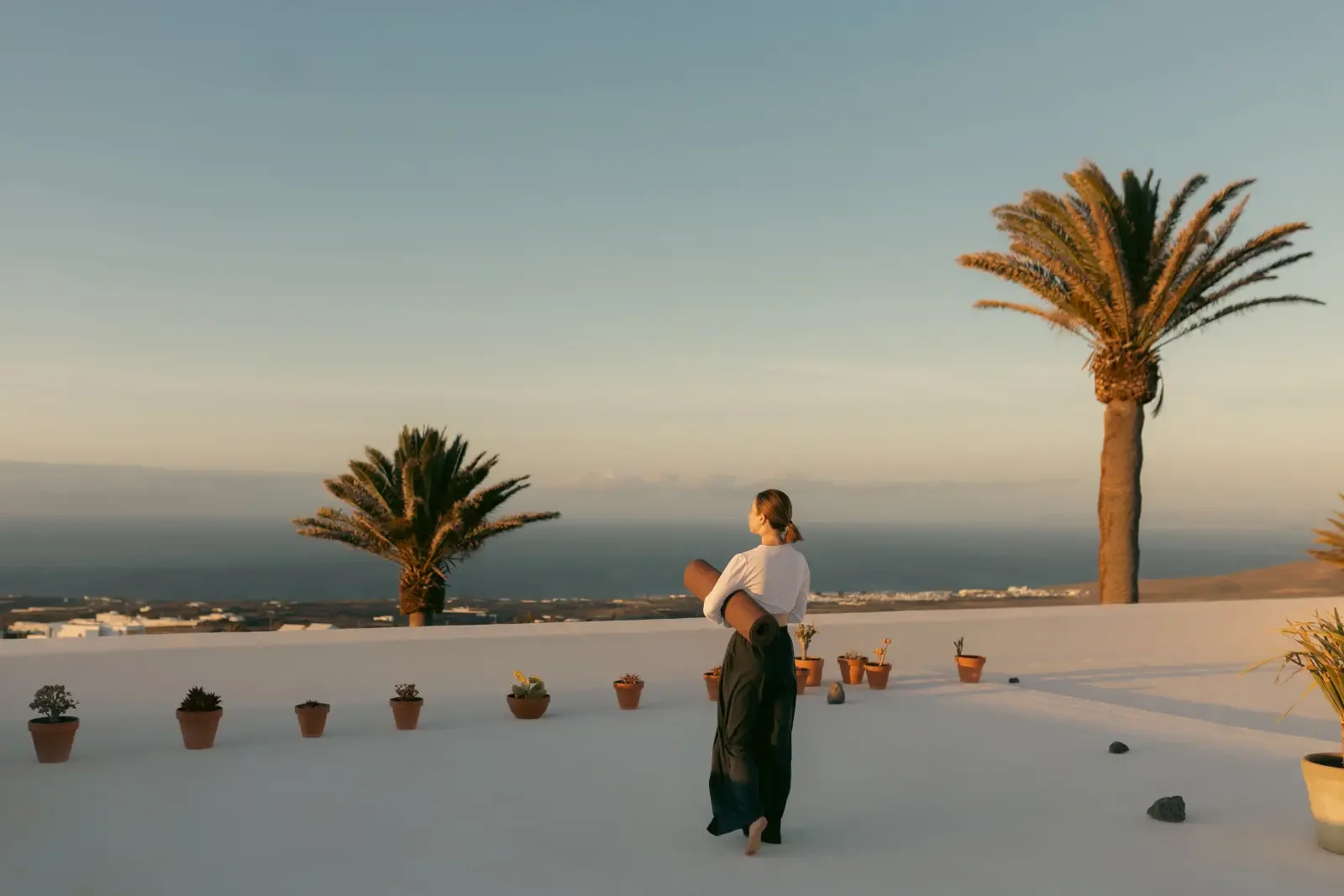A woman practicing yoga on a rooftop terrace with potted plants, palm trees, and a view of the ocean at sunset.