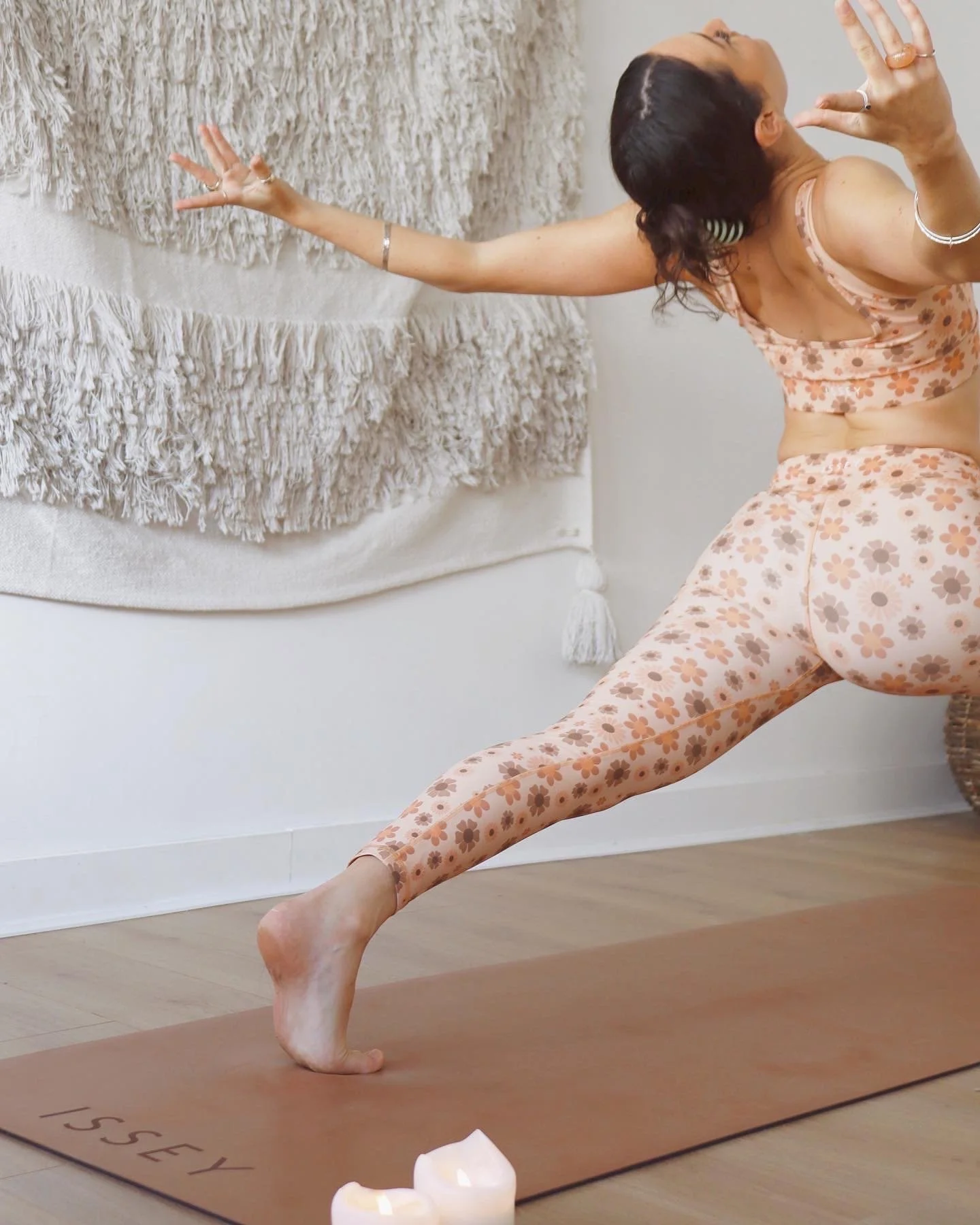 A woman practicing yoga indoors on a brown yoga mat with "ISSY" written on it, wearing floral-patterned activewear, stretching with arms extended and head turned back.
