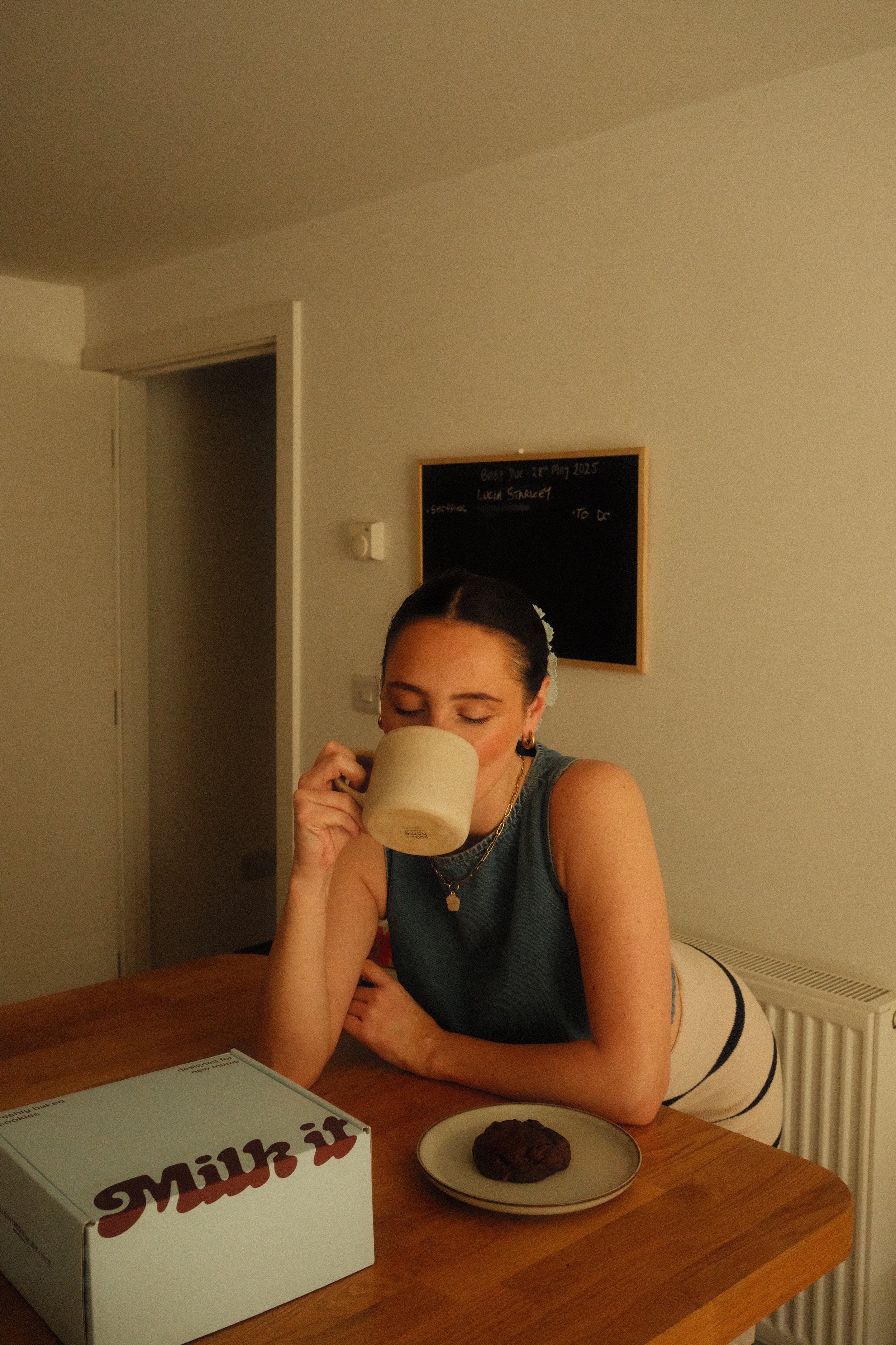 A woman with dark hair in a bun, wearing a sleeveless top and jewelry, is sitting at a wooden table, drinking from a beige mug with a cookie and a takeout box on the table.