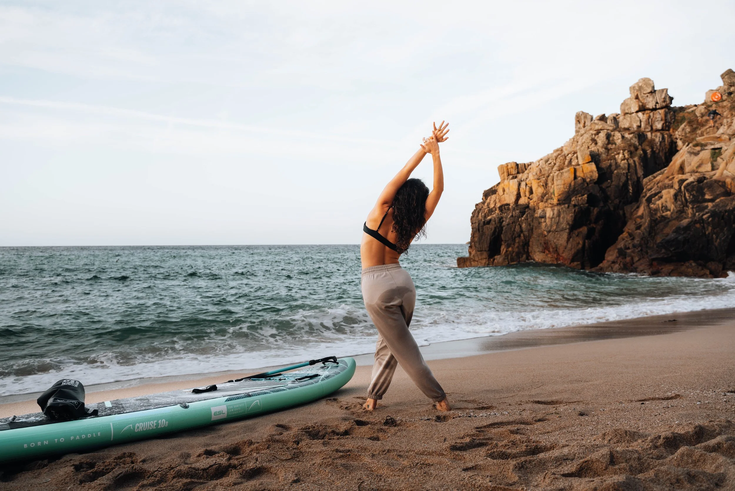 A woman practicing yoga on a beach near rocks with an ocean in the background, and a paddleboard lying on the sand nearby.