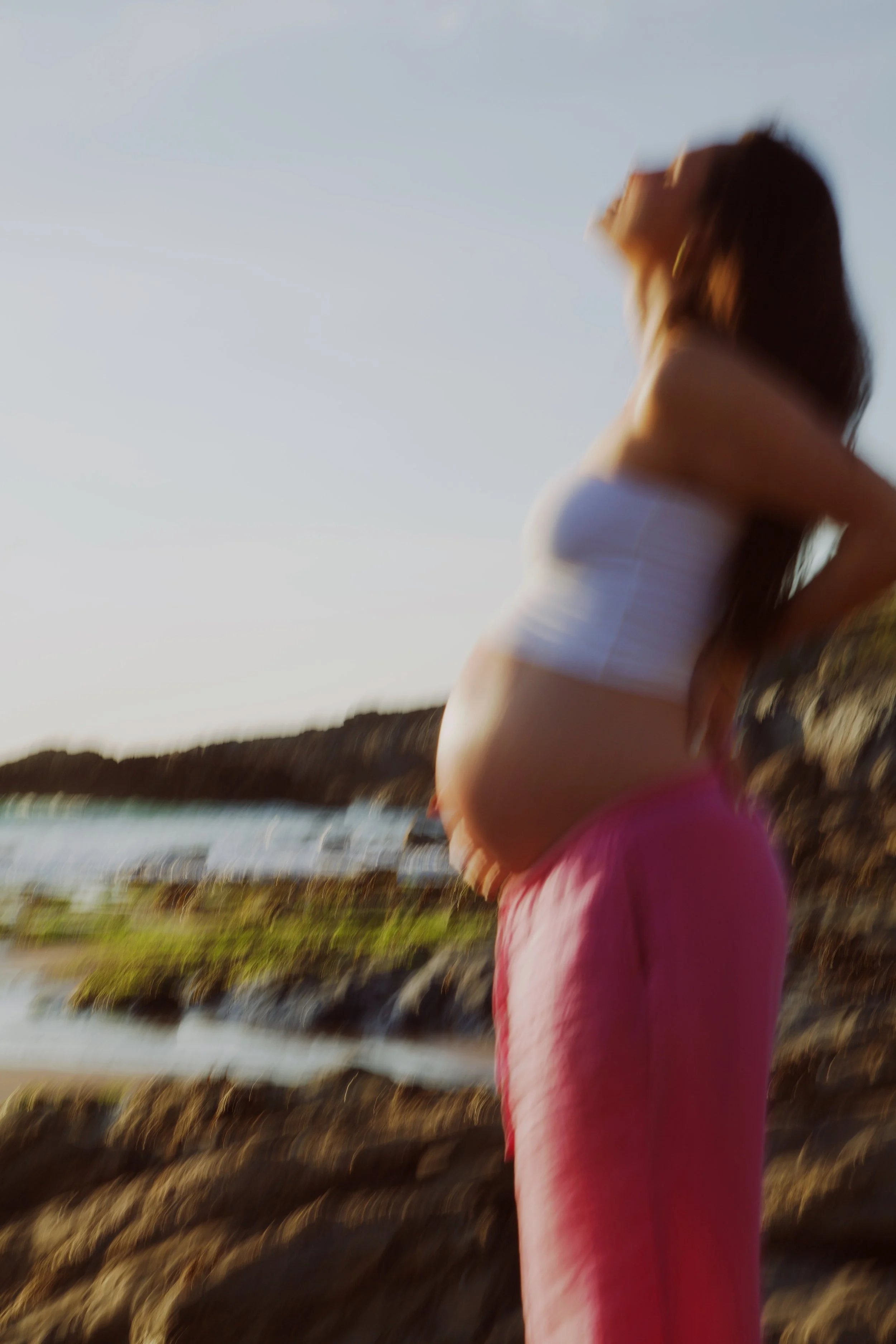 A pregnant woman standing outdoors near the water, with her hand on her belly, during sunset or late afternoon.