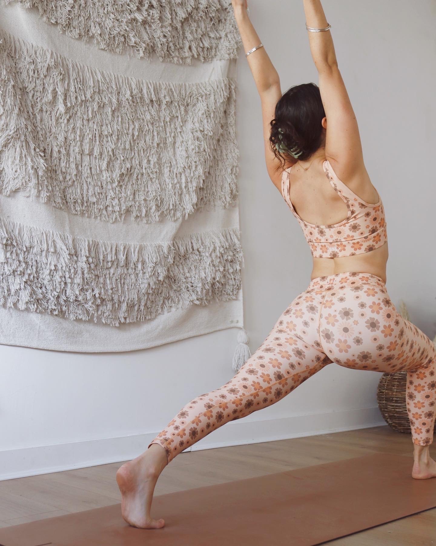 A woman practices yoga in a lunge pose on a yoga mat indoors, wearing floral leggings and a matching sports bra, with a textured wall hanging in the background.