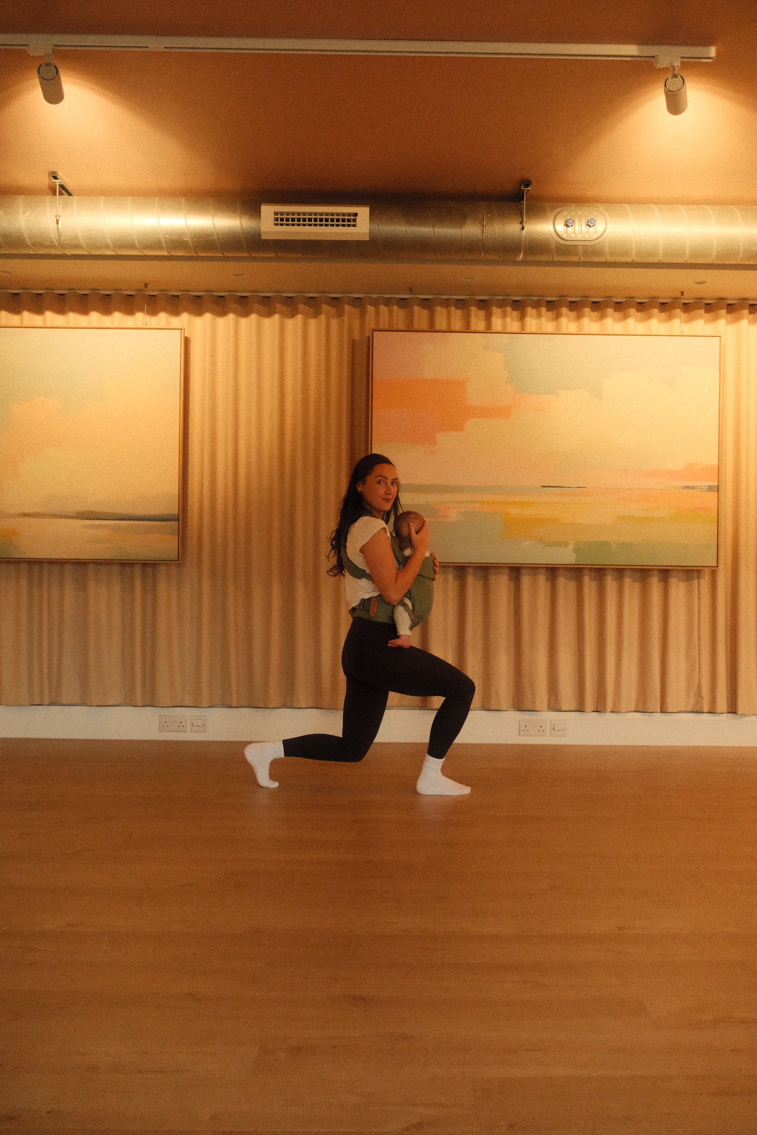 A woman in workout clothes holding a baby while doing a lunge exercise in a room with wooden flooring and pastel-colored abstract paintings on the wall.
