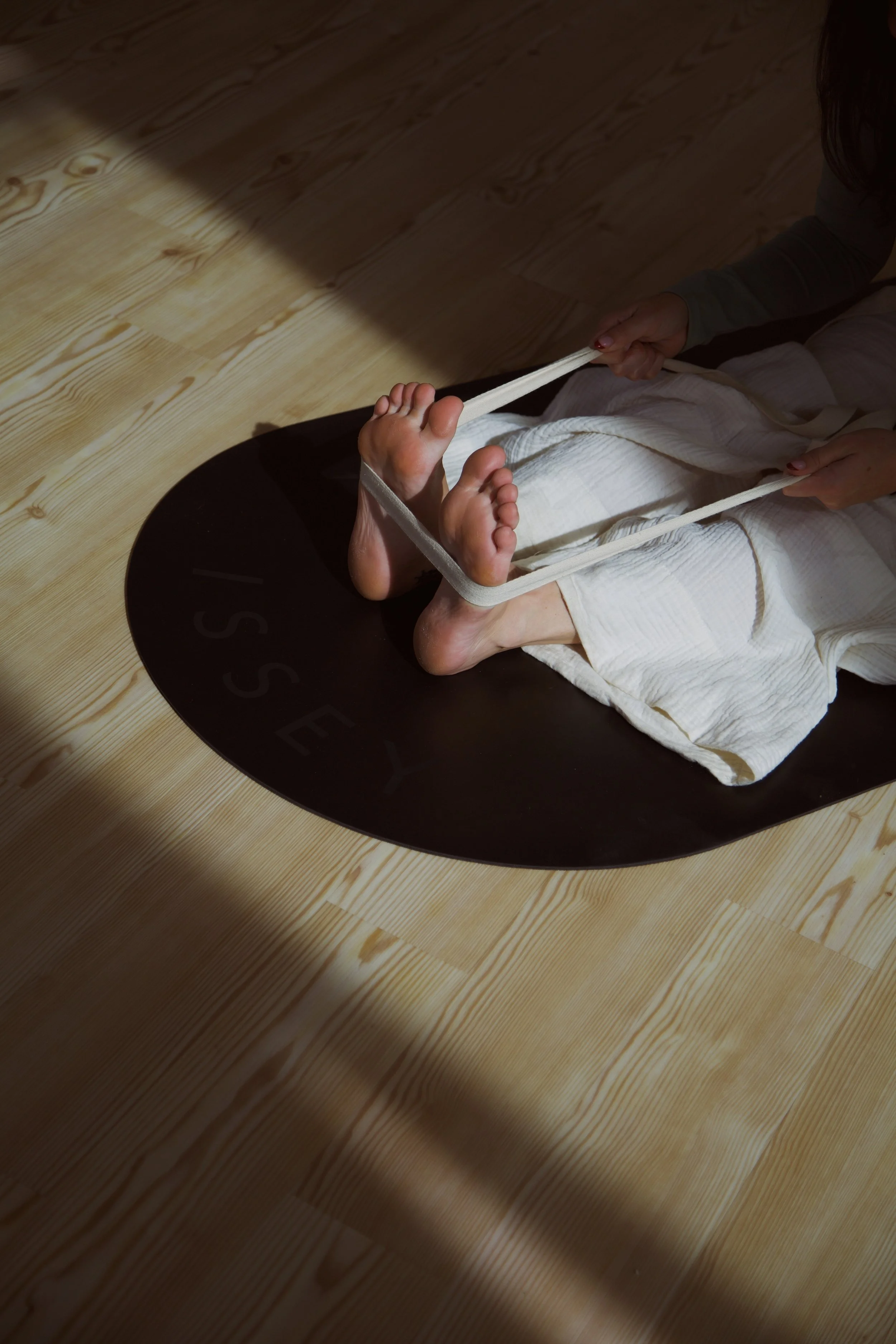 Person in a white karate uniform sitting on a black mat, holding a person-sized resistance band with their feet. The mat has the word ''SSEY'' partially visible.