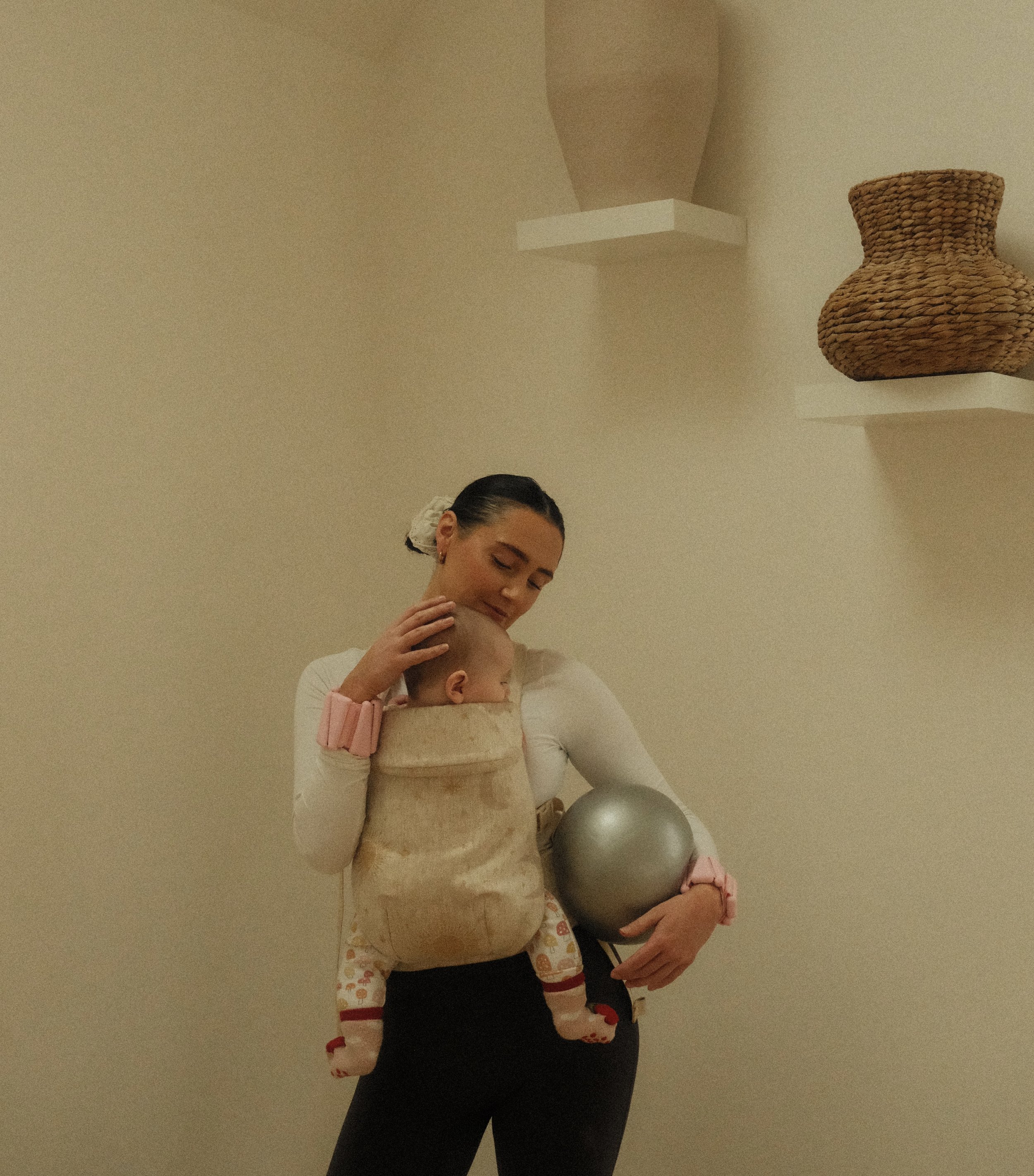 Woman carrying a baby in a beige carrier on her chest, holding a silver exercise ball, standing against a plain wall with decorative shelves.