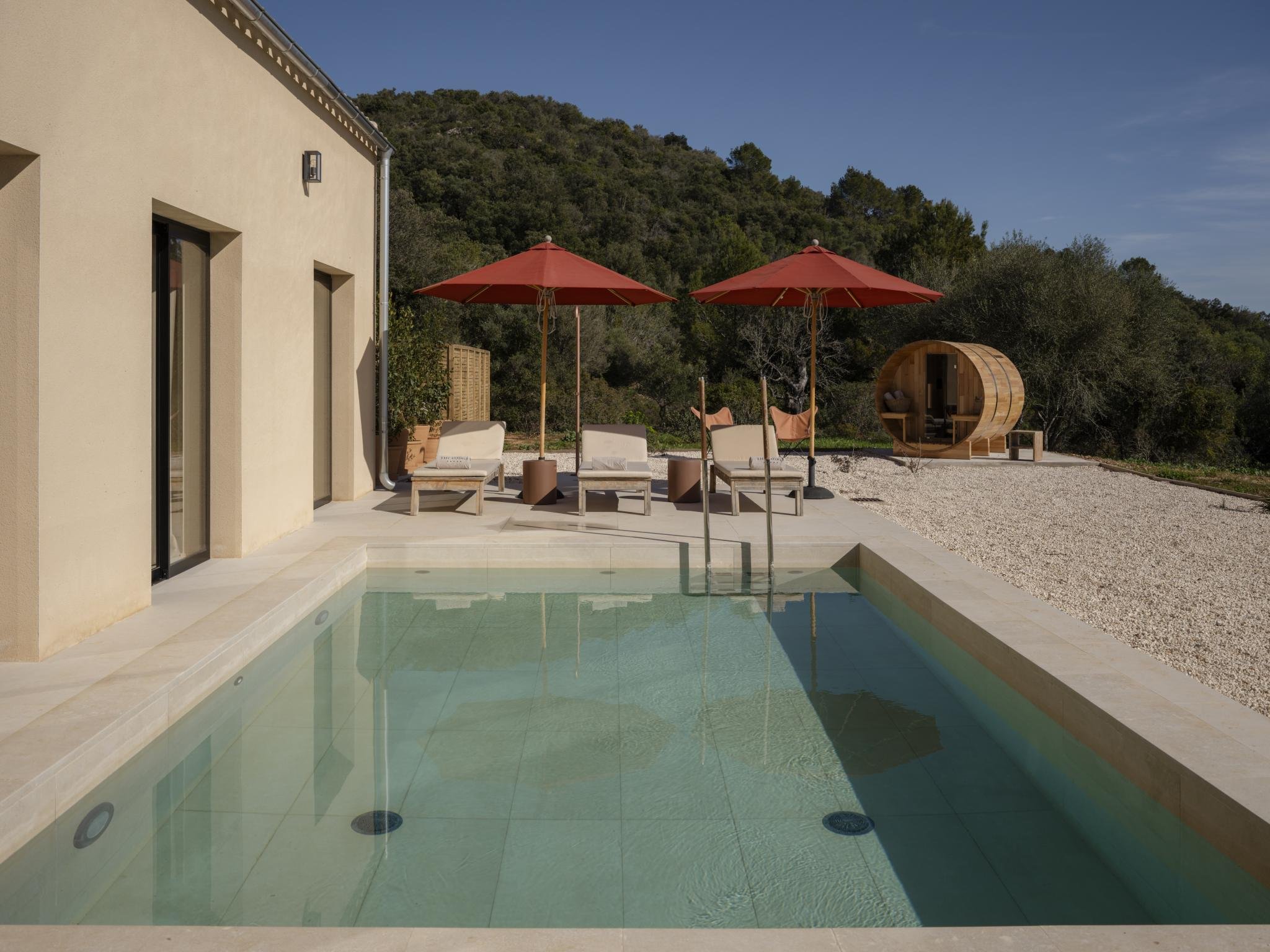 Modern backyard with a small swimming pool, two red umbrellas, lounge chairs, a wooden cabana, and a garden backdrop.