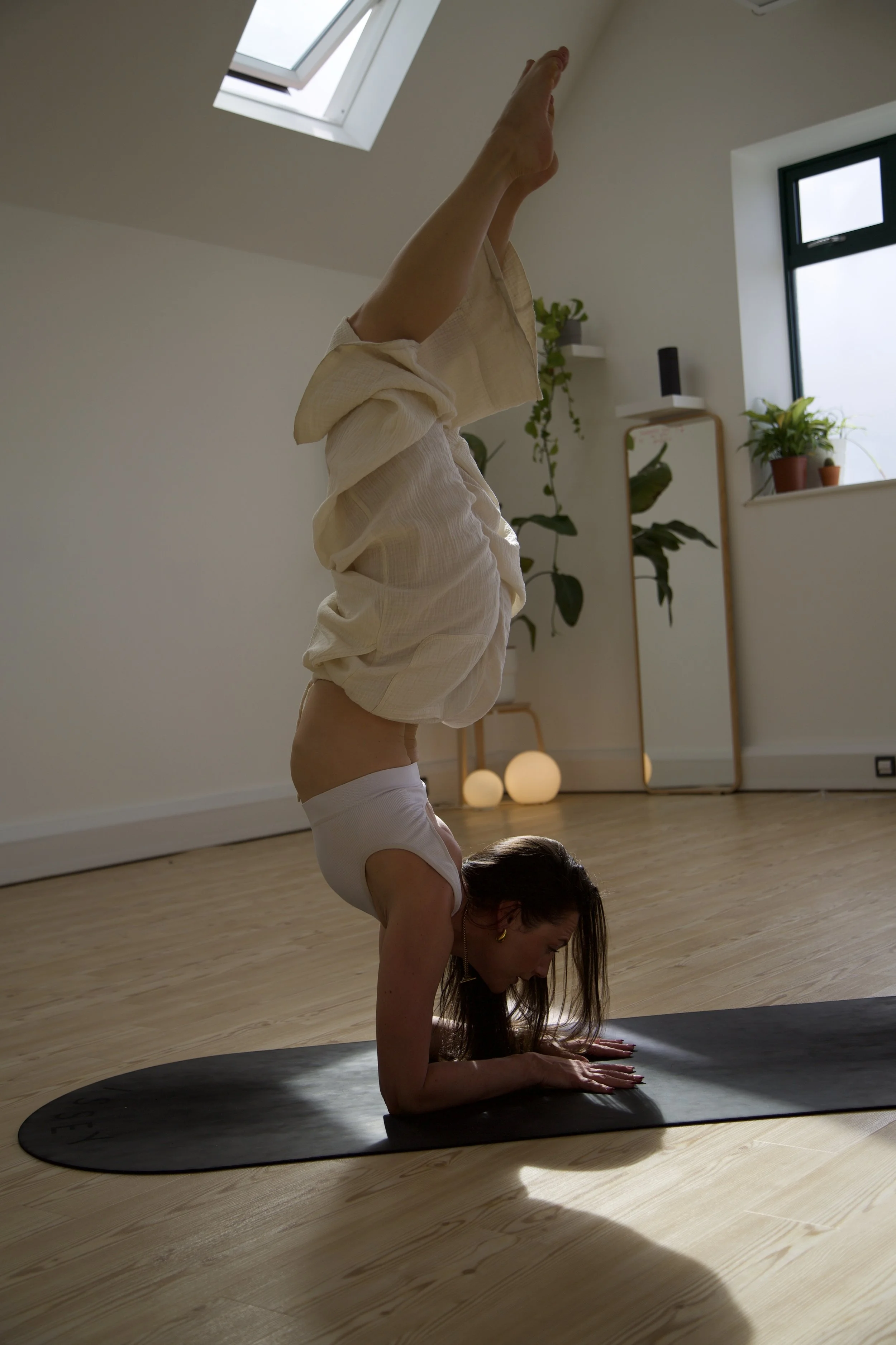 A woman practicing yoga indoors, balancing on her forearms with her legs extended upward in a headstand pose on a black yoga mat.