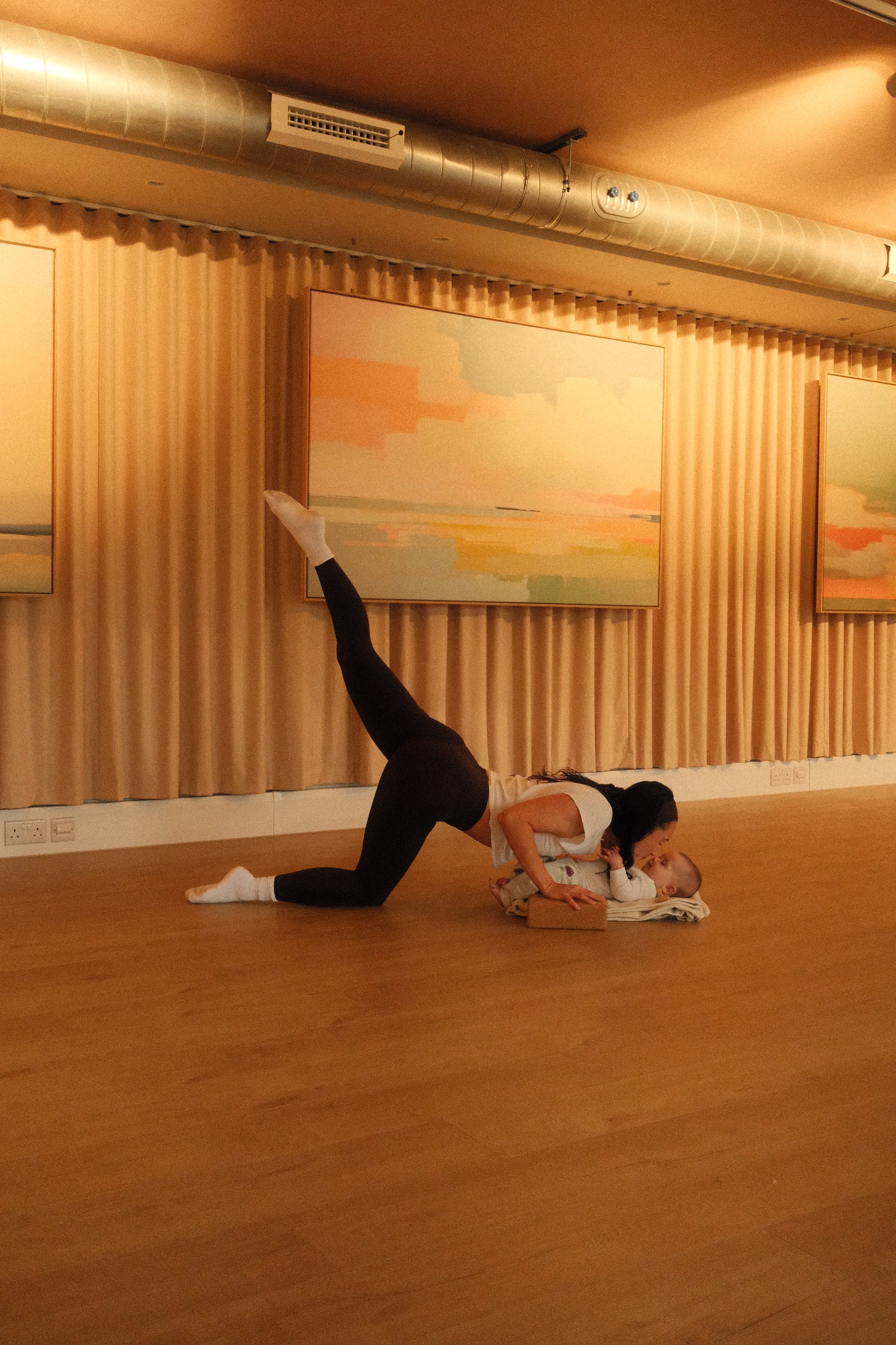 A woman practicing yoga with a baby lying on a mat in a dance studio with artwork on the walls.