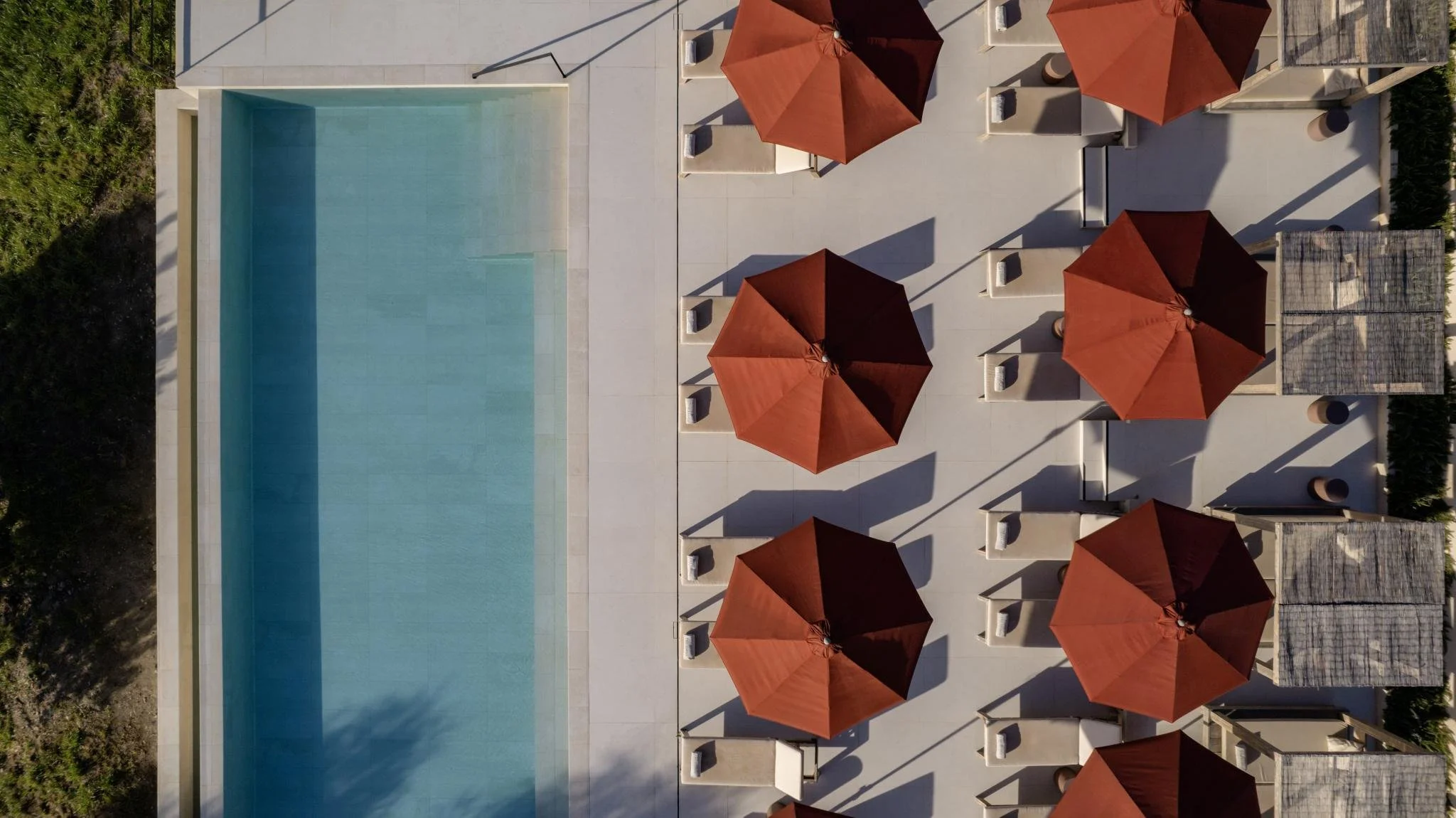 Top-down aerial view of a swimming pool with a light teal interior, surrounded by a white tiled deck. There are six large rust-colored umbrellas providing shade, each with lounge chairs underneath. Small tables and additional lounge chairs are also visible around the umbrellas.