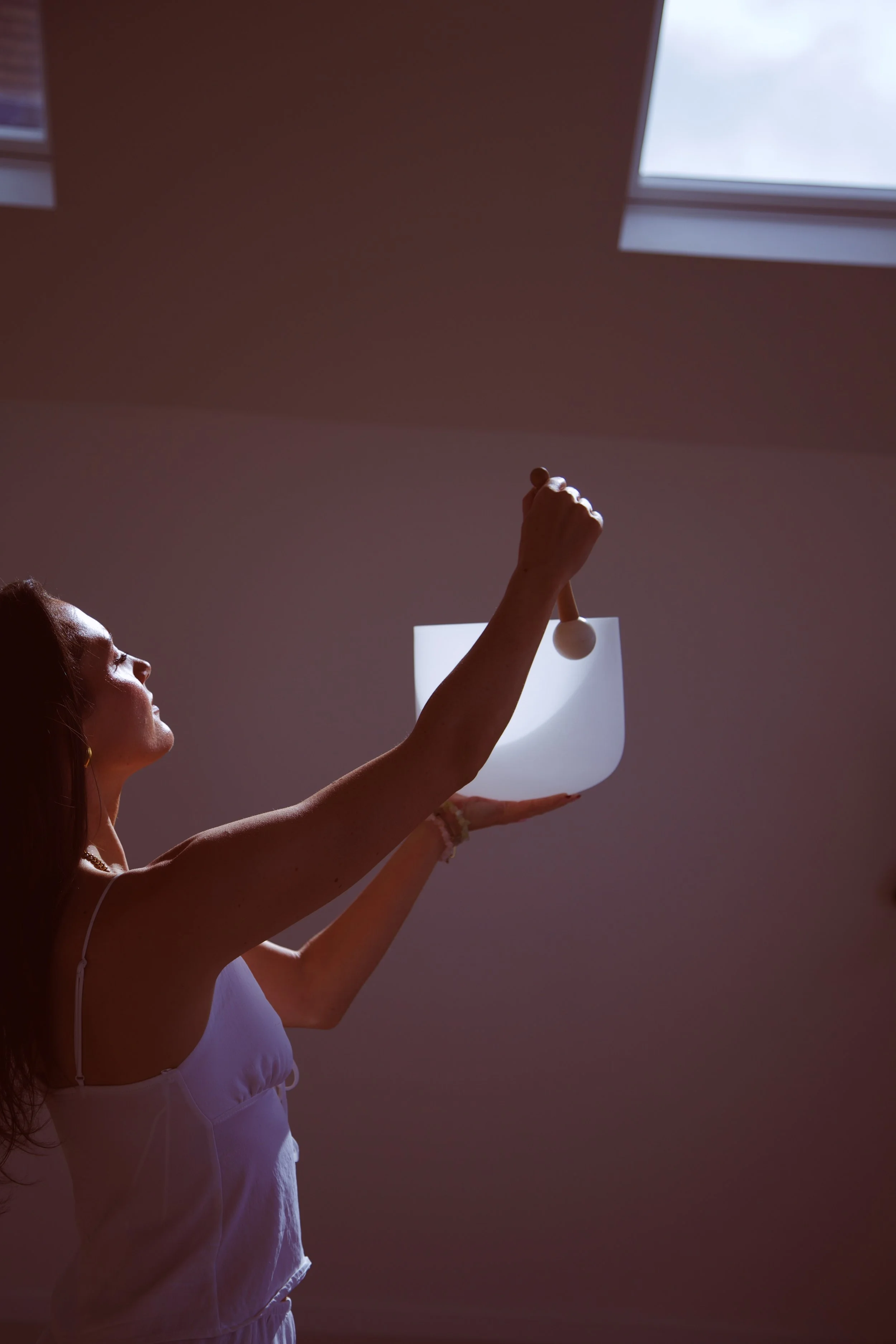A woman holding a singing bowl and a mallet in a room with a window in the background.