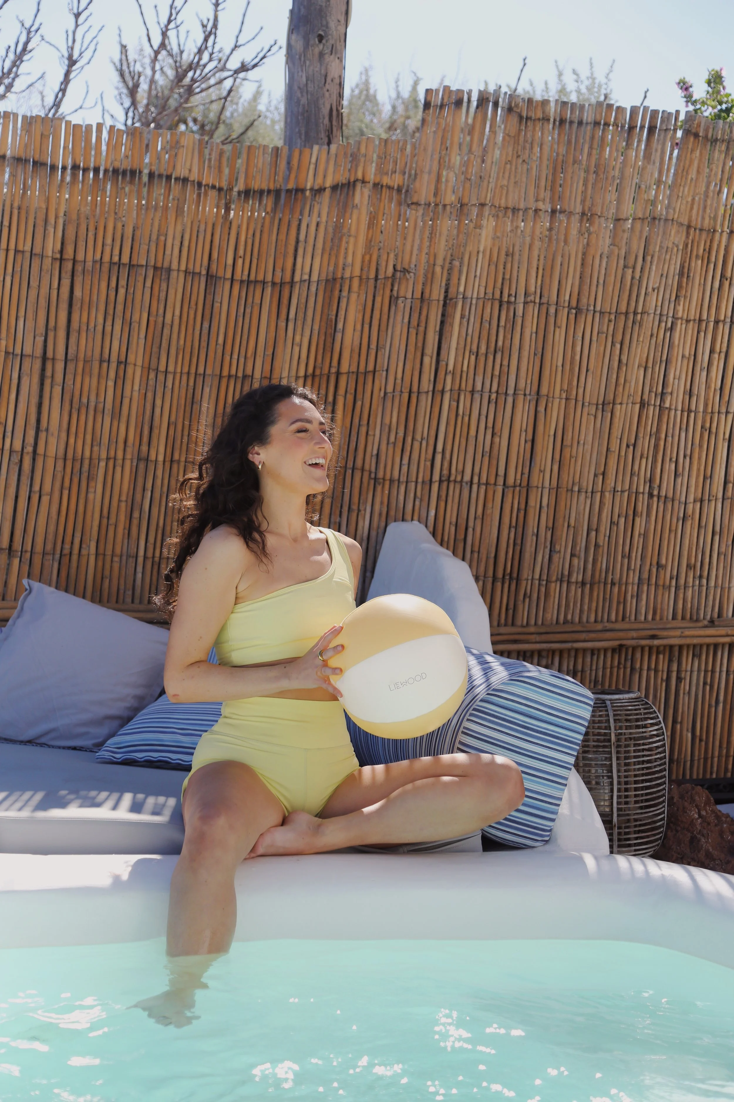 Woman sitting by the pool holding a yellow and white beach ball, smiling and wearing a yellow swimsuit, with a bamboo fence in the background.