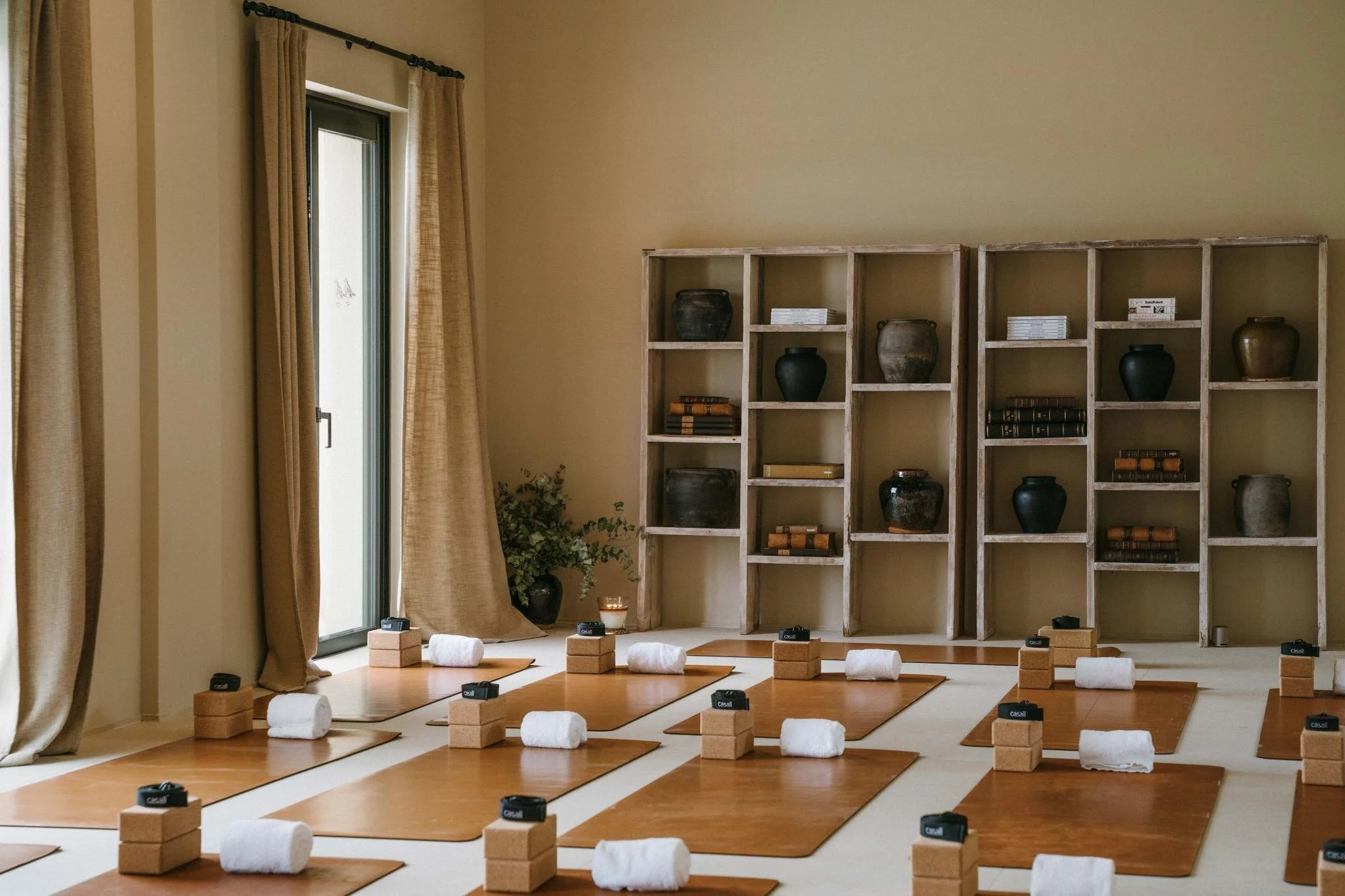 Yoga mats with rolled towels, blocks, and eye pillows arranged in a room with large windows and wooden shelves with vases and books.